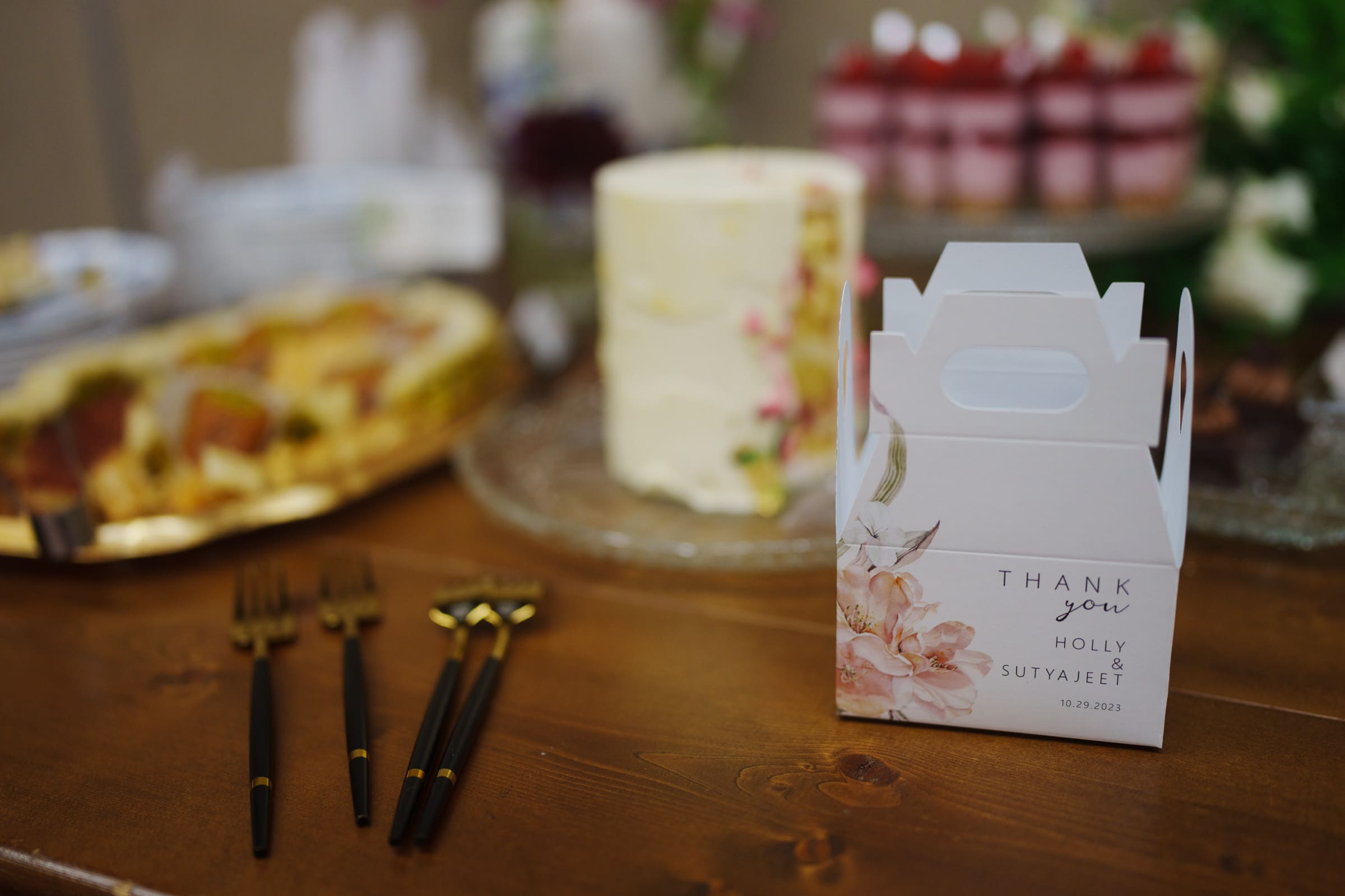 A wedding table at the JC Raulston Arboretum showcases a white gift box adorned with floral designs and "Thank You, Holly & Sutvajeet, 10-20-2023" print. In the background, there's a white cake and various desserts, with black and gold utensils neatly arranged nearby.
