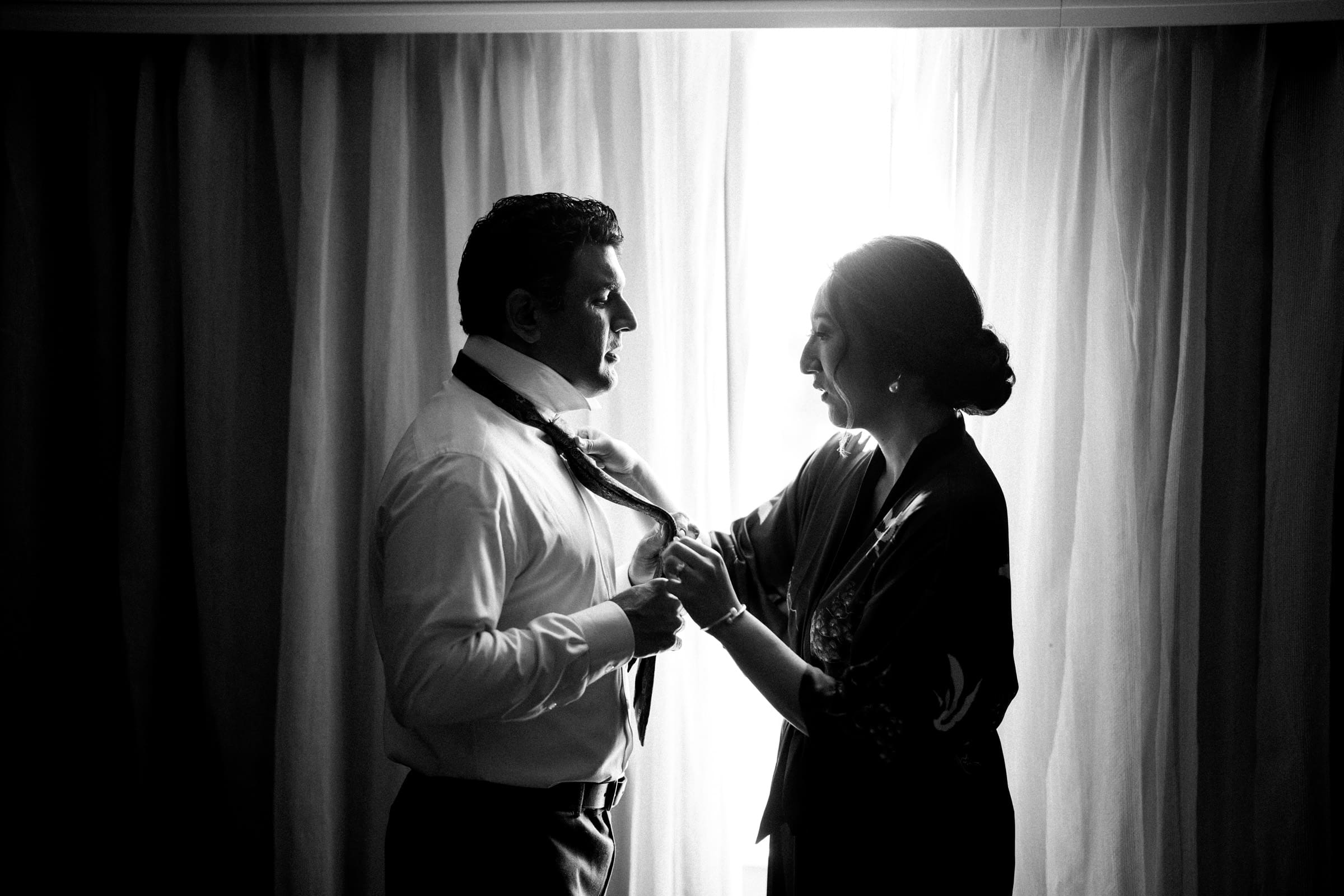 A black and white image captures Holly helping Sut with his tie, both elegantly dressed for their JC Raulston Arboretum wedding. Standing in front of a window with drawn curtains, the light casts a striking silhouette of the couple's intimate moment.