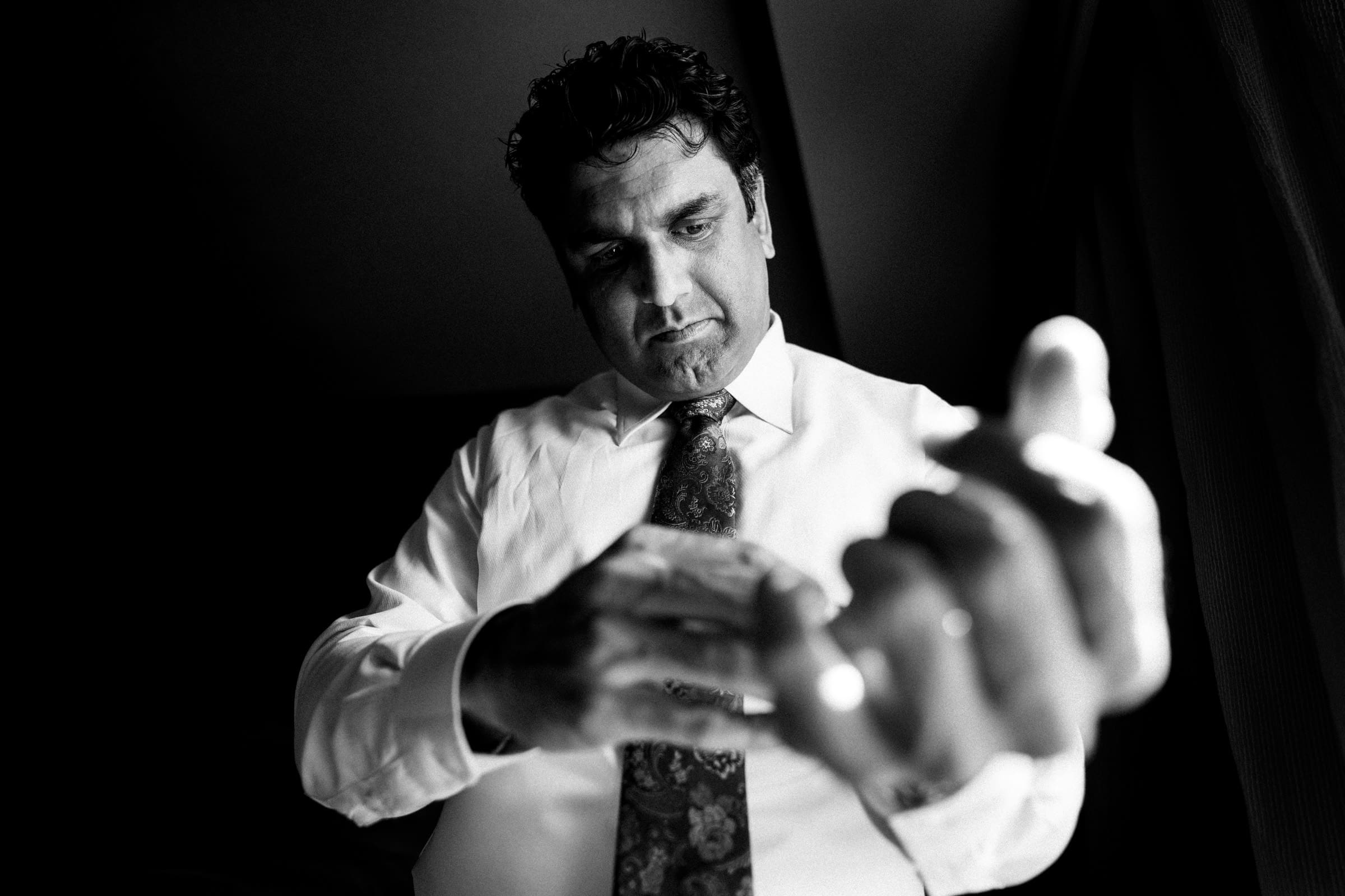 In a black and white image, a man in a white shirt and patterned tie adjusts his sleeve with meticulous care. The lighting casts deep shadows, emphasizing his focused expression, reminiscent of the elegant moments captured at JC Raulston Arboretum weddings like Holly & Sut’s.