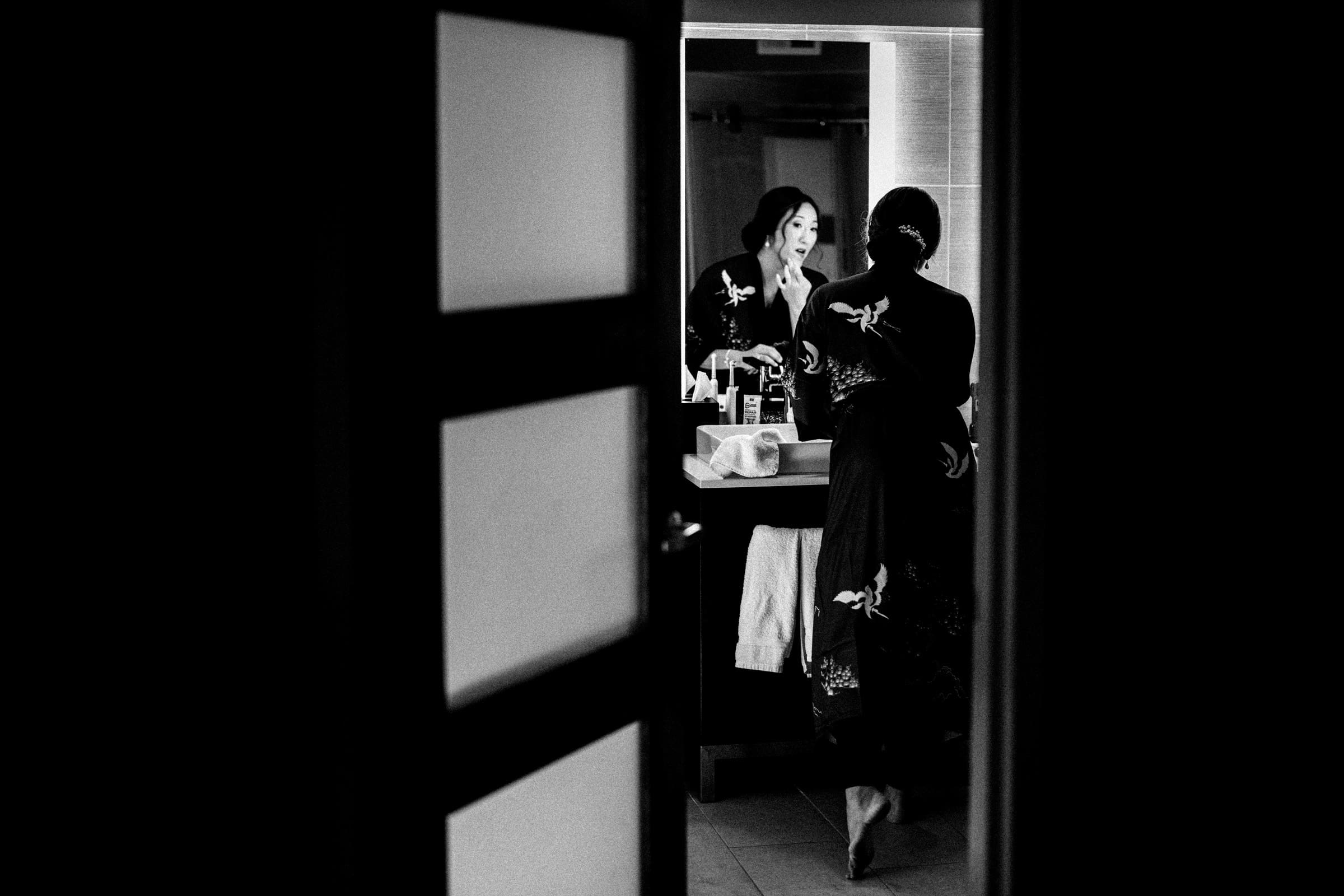 A black and white image captures Holly in a kimono, standing before a mirror, applying makeup for her JC Raulston Arboretum wedding with Sut. The scene, viewed through a partially open door, exudes intimacy and focuses on her serene reflection.