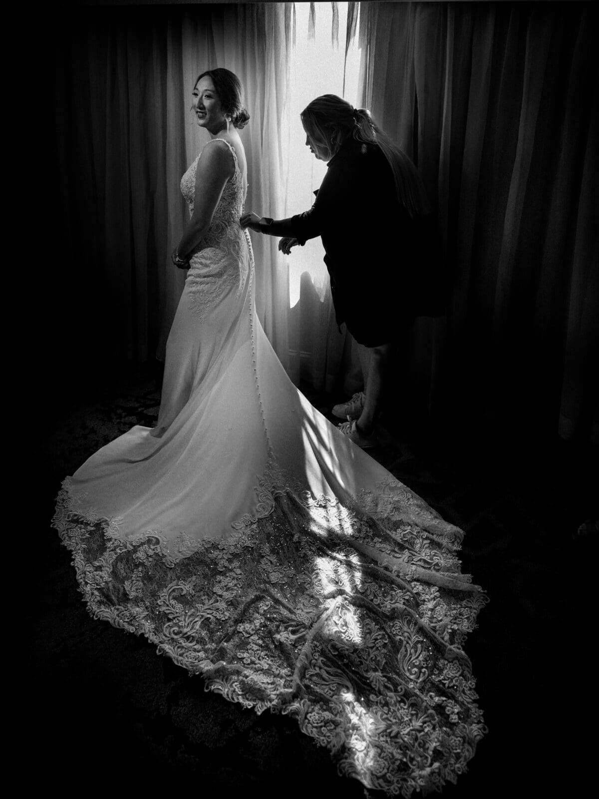 A bride in a lace wedding gown stands by a window at the JC Raulston Arboretum, while another person helps her with the dress. Sunlight filters through the curtains, illuminating the elegant train at Holly and Sut's wedding.