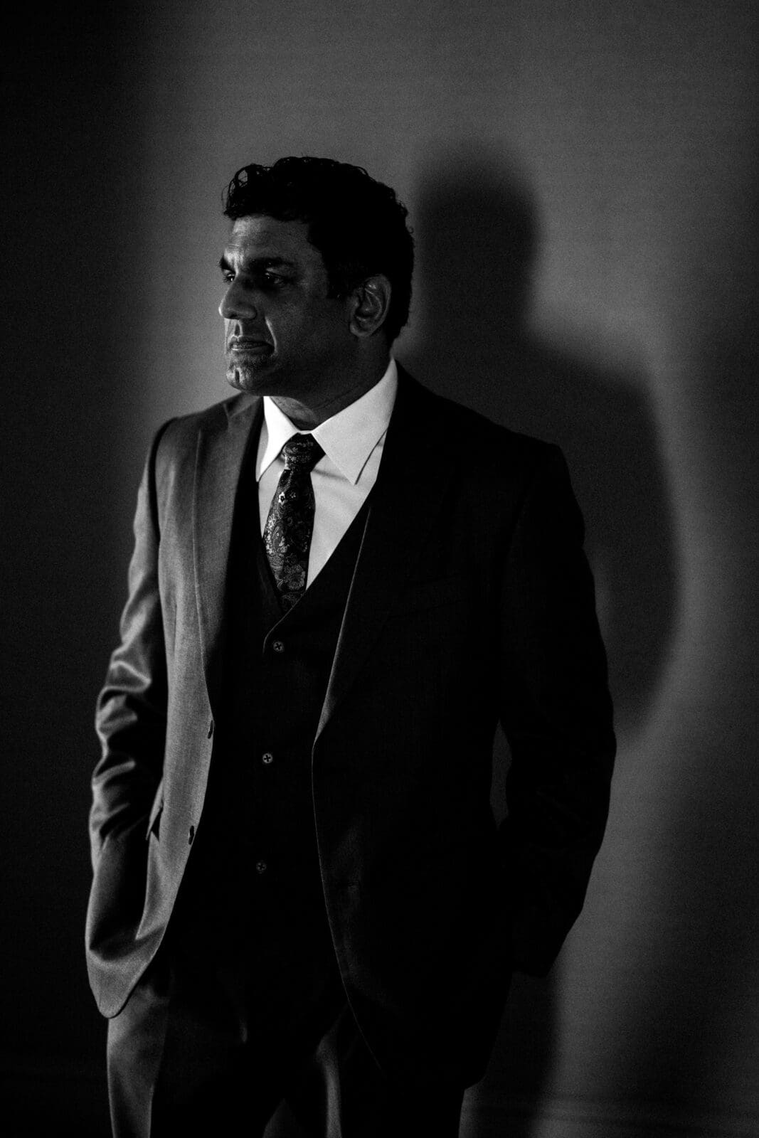 In a dimly lit room, a black and white image captures a person in a suit and tie, hands in pockets. Their shadow stretches onto the wall, evoking contemplation—a moment reminiscent of quiet reflections at Holly & Sut's JC Raulston Arboretum wedding.