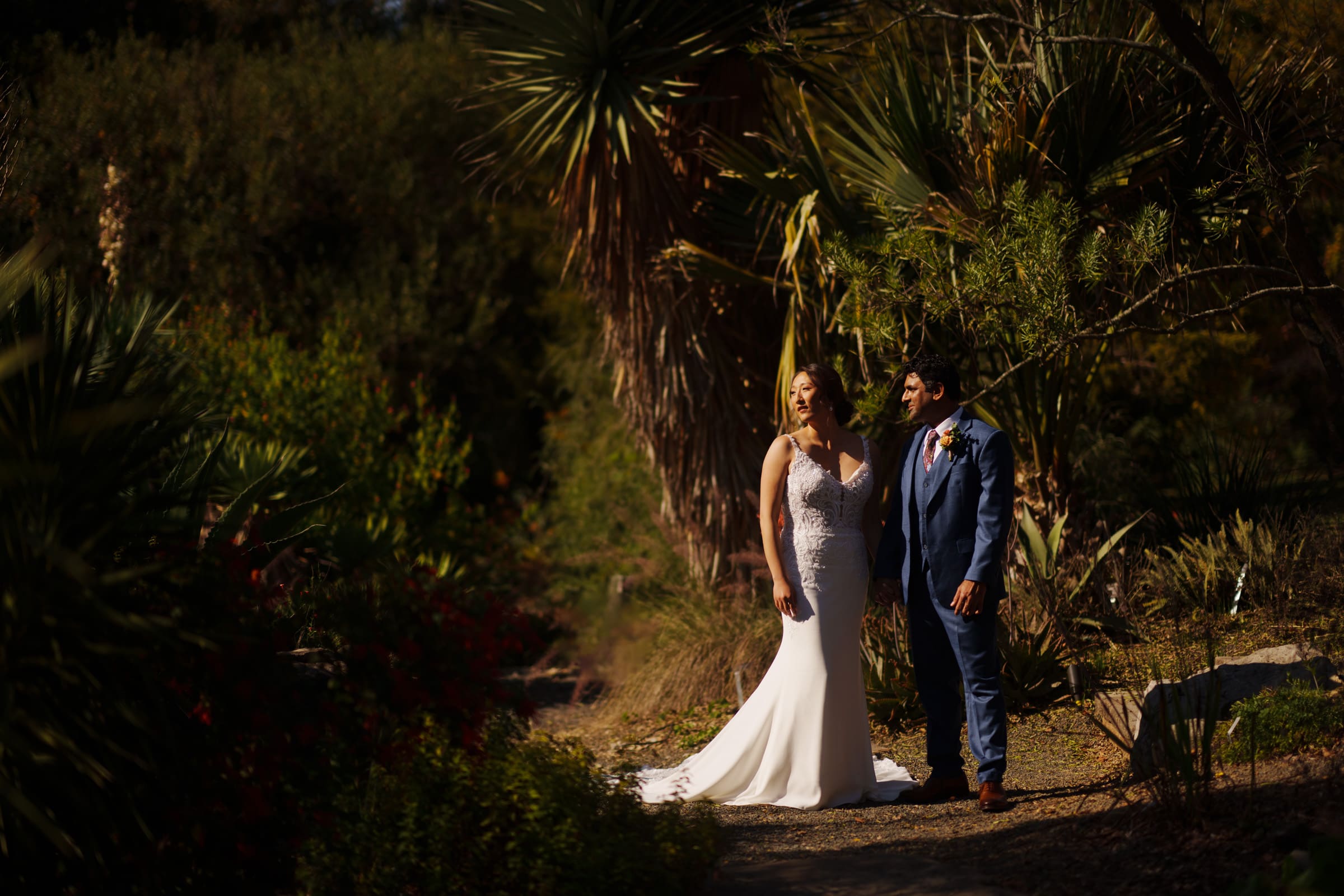 In the sunlit JC Raulston Arboretum, Holly and Sut stand serenely amidst lush greenery and towering palm trees. She glows in her white wedding dress, while he looks sharp in a blue suit, both embracing the peaceful beauty of their special day.
