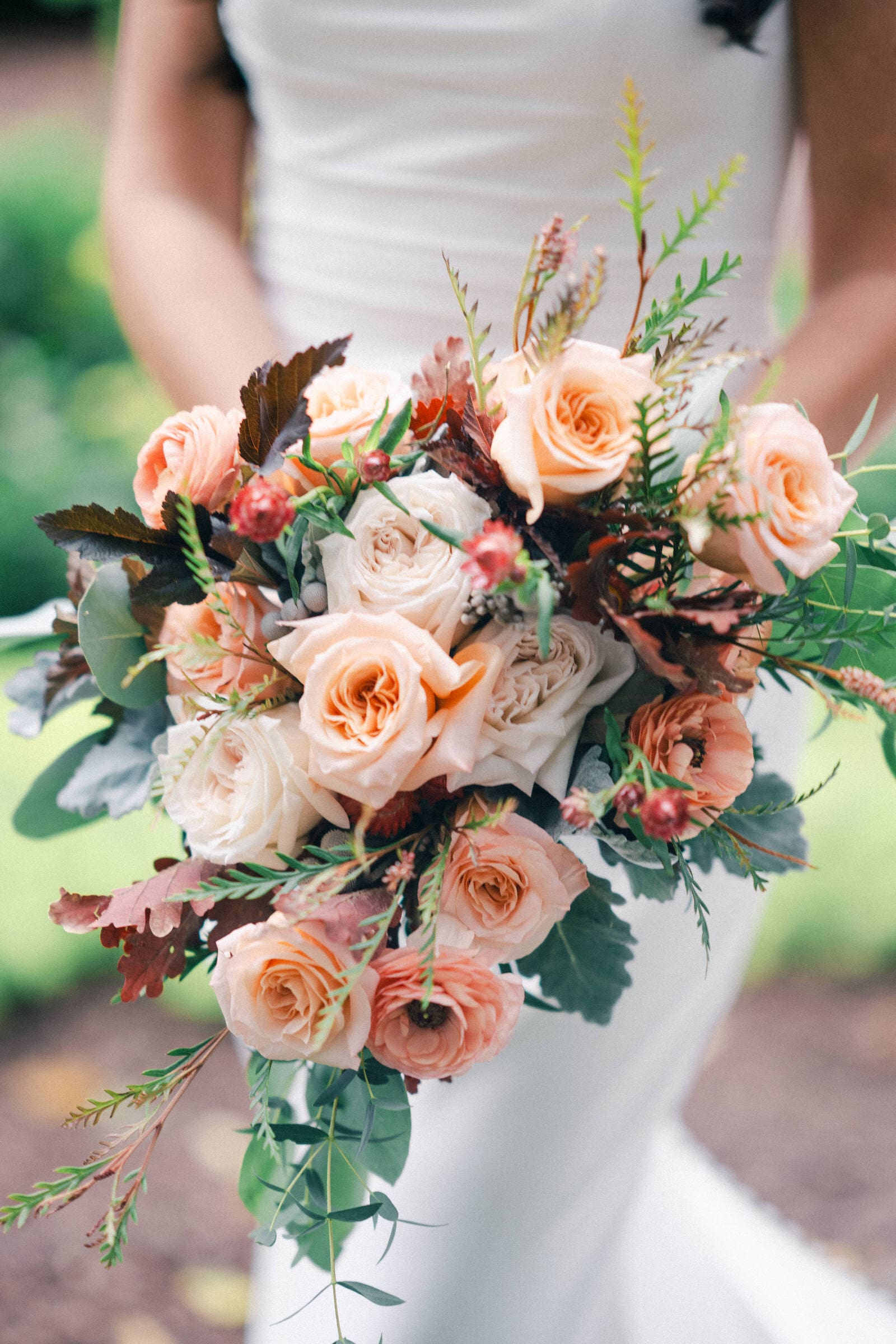 At the picturesque Rose Hill Estate wedding, a bride holds a bouquet of peach and white roses mixed with green leaves and brown accents, set against a blurred natural background.
