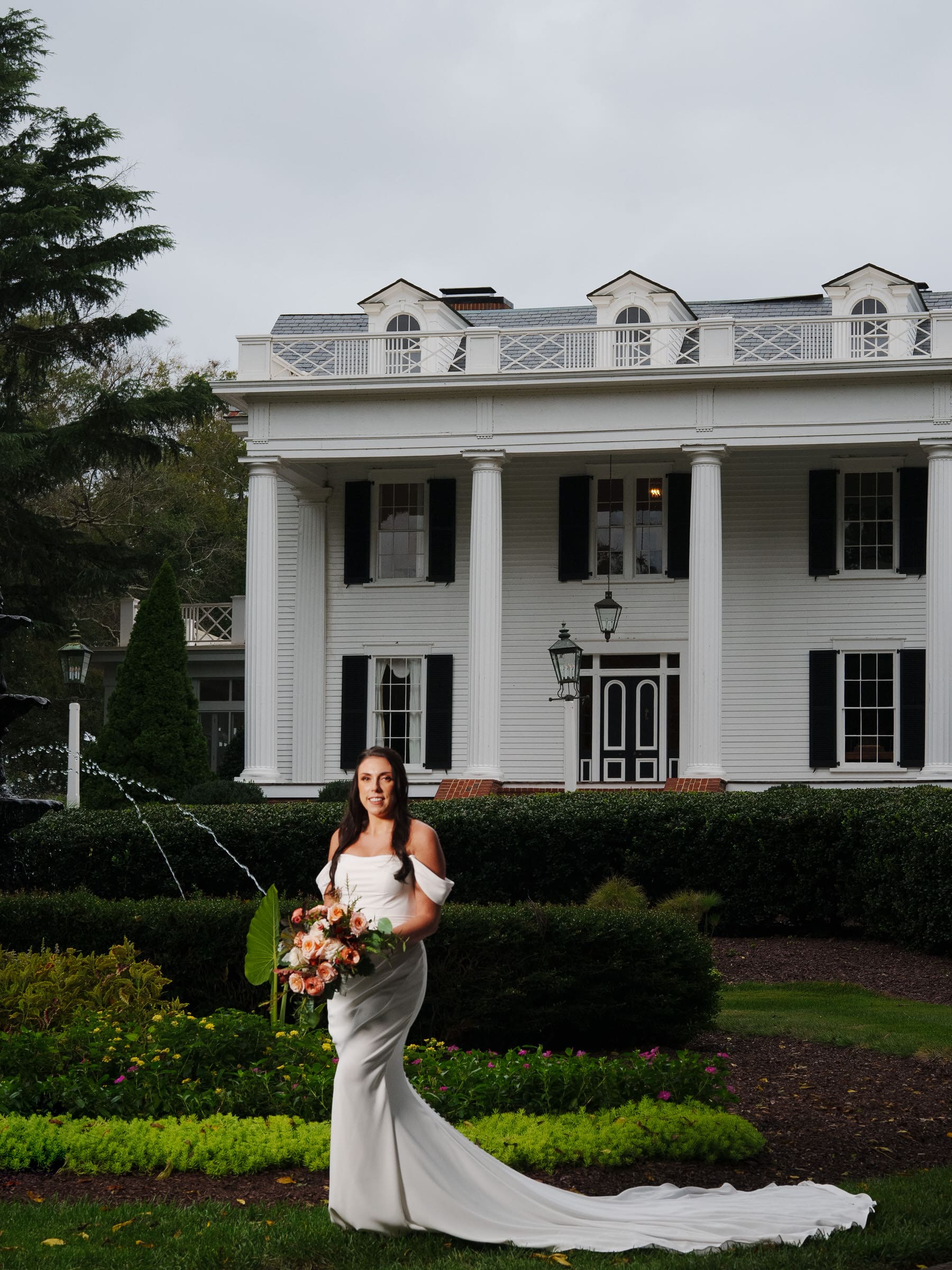 A bride in a white gown stands holding a bouquet in front of Rose Hill Estate, a large colonial-style mansion with green shutters and columns. The garden, perfect for a wedding, features a small fountain and neatly trimmed hedges.