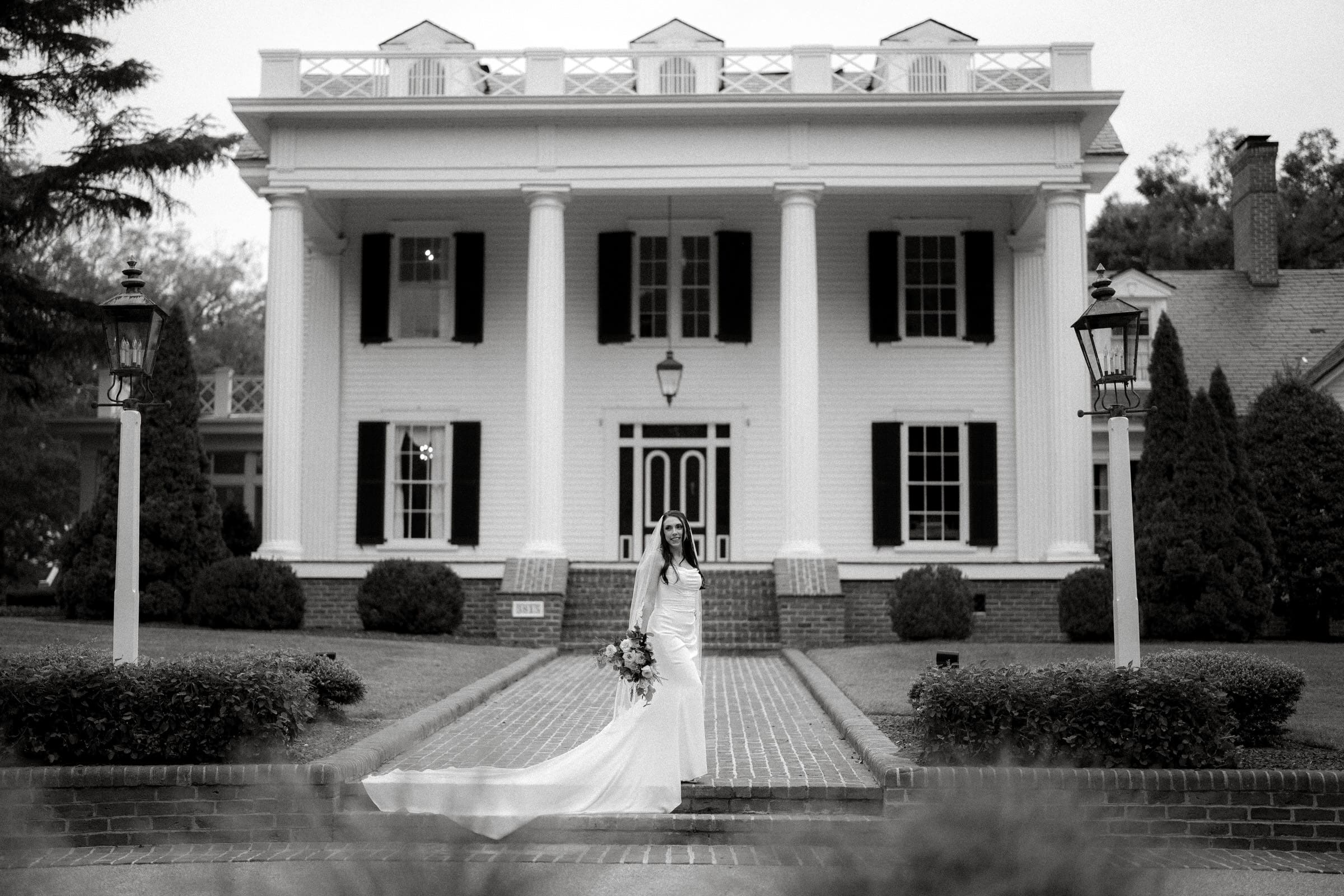 A bride stands in front of the grand, white-columned Rose Hill Estate, holding a bouquet. She wears a long dress with a flowing train that cascades behind her. The mansion features large windows with shutters and a lantern-lit path leading to the elegant entrance of this stunning wedding venue.
