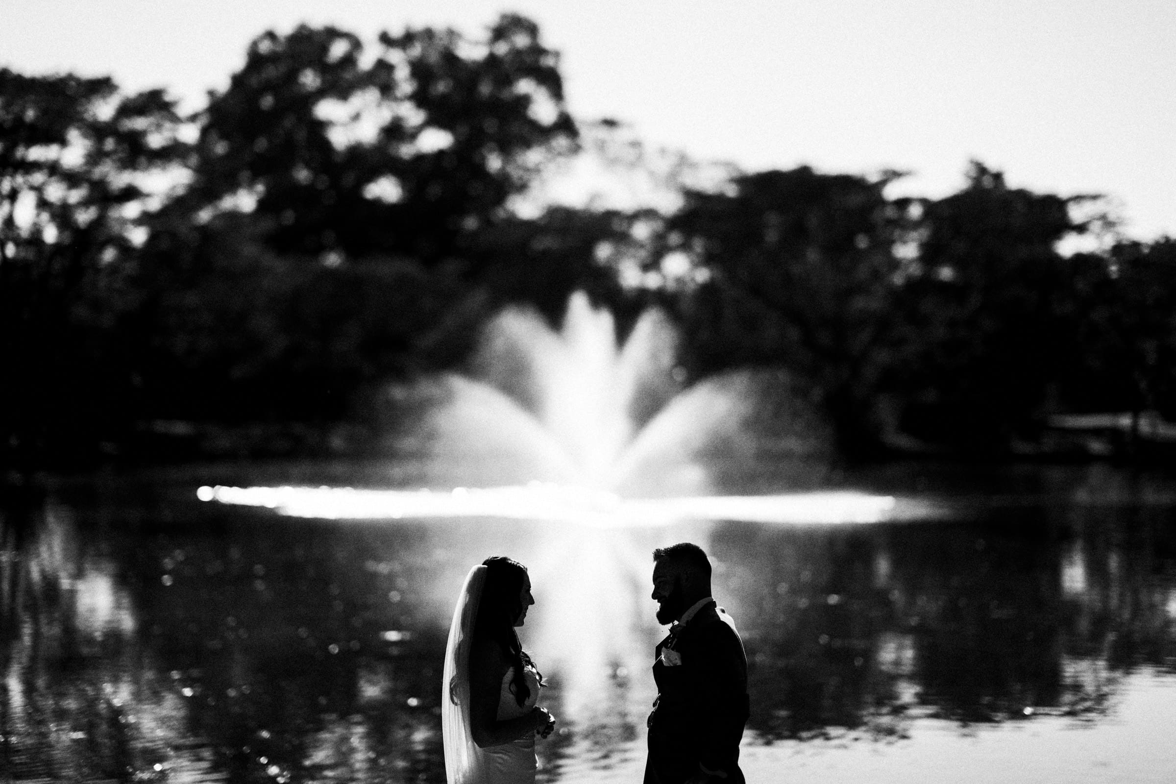 A silhouetted couple stands by a serene lake at Rose Hill Estate, with a fountain in the background. The scene is framed by trees, and the sunlight creates a peaceful atmosphere, perfect for an unforgettable wedding moment.