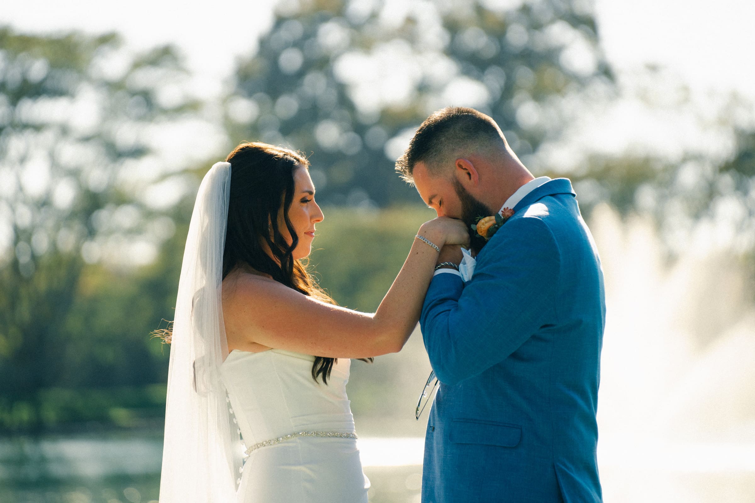 At a charming Rose Hill Estate wedding, a bride and groom stand facing each other outdoors. The groom, in a blue suit, gently kisses the bride's hand. She is elegantly dressed in a white gown with a veil. They are by a body of water with a picturesque fountain and lush trees in the background.