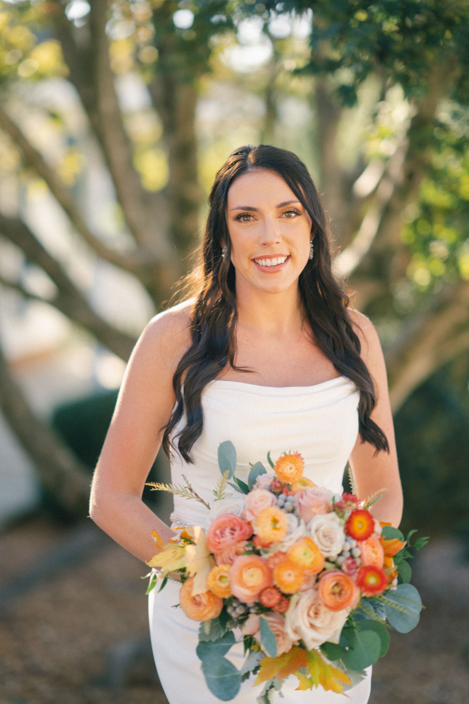 A bride with long brown hair stands outdoors at Rose Hill Estate, holding a colorful bouquet of flowers. She's wearing a white wedding dress, with green foliage and soft sunlight filtering through the trees in the background.