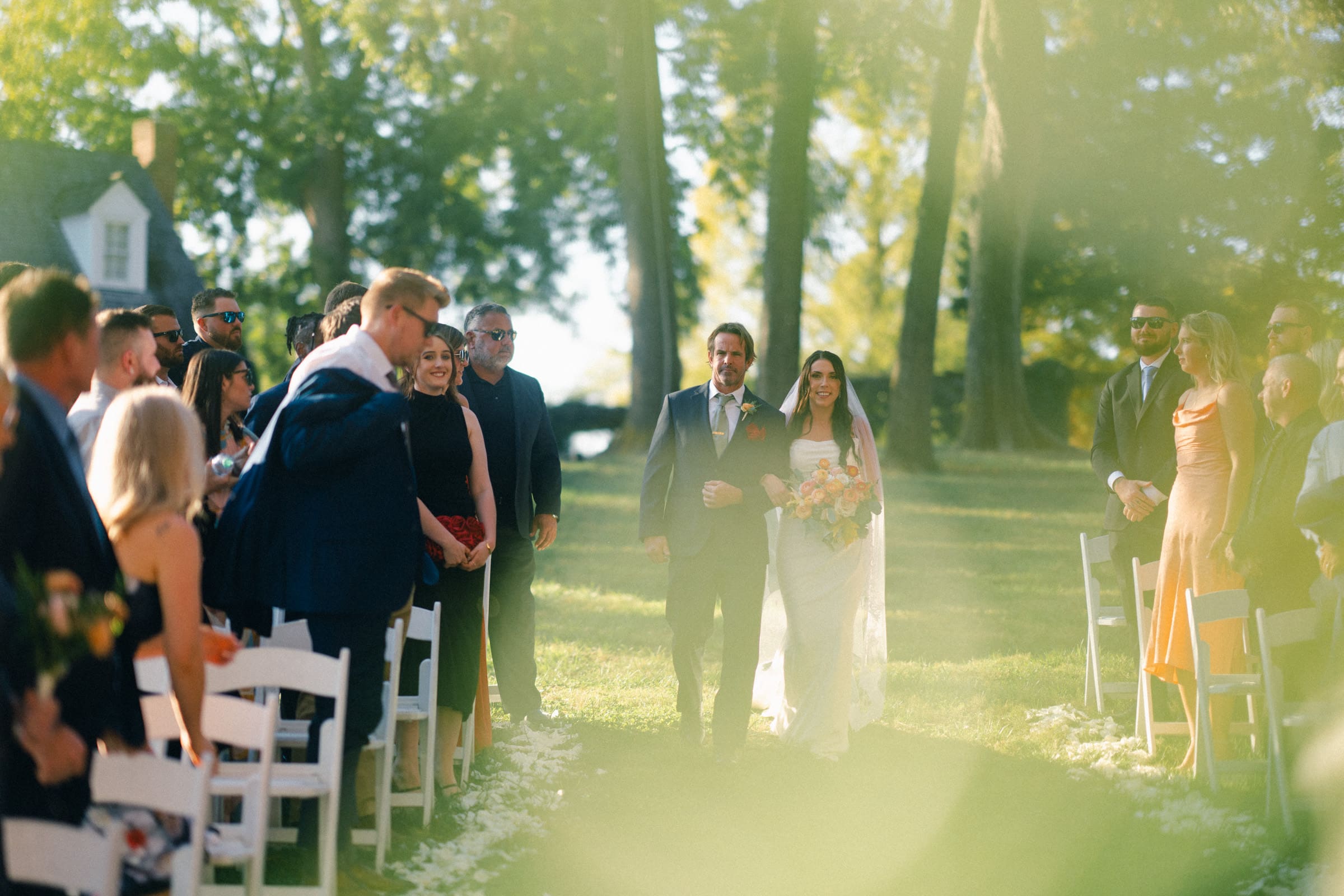 At a picturesque Rose Hill Estate wedding, a bride in a white dress and a groom in a suit walk down an outdoor aisle surrounded by guests seated on white chairs. Sunlight filters through trees, creating a dreamy atmosphere as guests watch and smile while the couple passes.