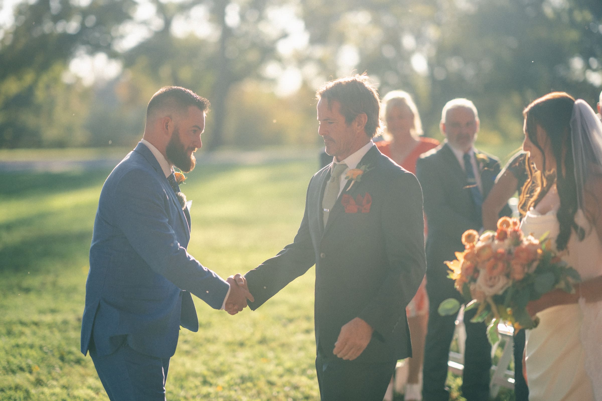 At the sunny Rose Hill Estate wedding, a groom in a blue suit shakes hands with the father of the bride. The bride stands nearby, holding a bouquet on the grassy lawn as guests enjoy this beautiful outdoor ceremony in the background.