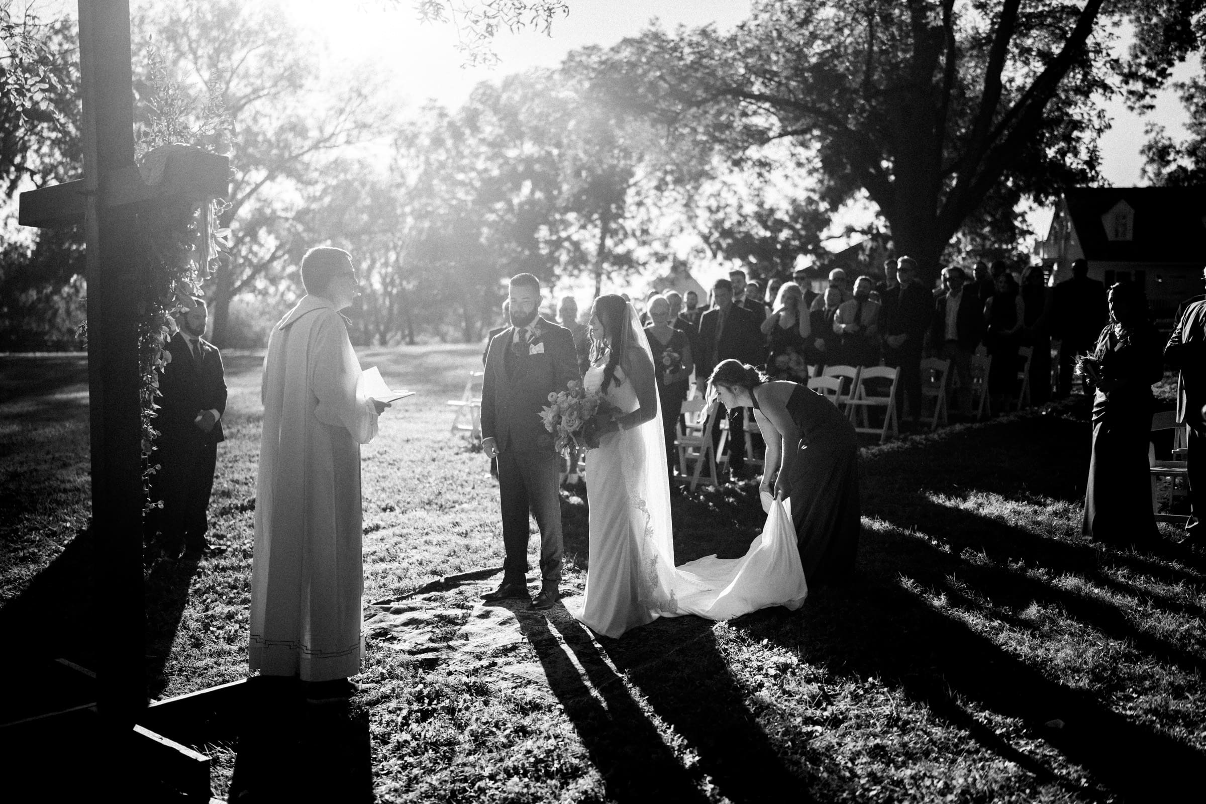 A black and white image of a Rose Hill Estate wedding captures an outdoor ceremony. A couple stands facing a person in ceremonial attire, with the bride adjusting her gown. Guests are seated nearby under tall trees, sunlight streaming through the branches.