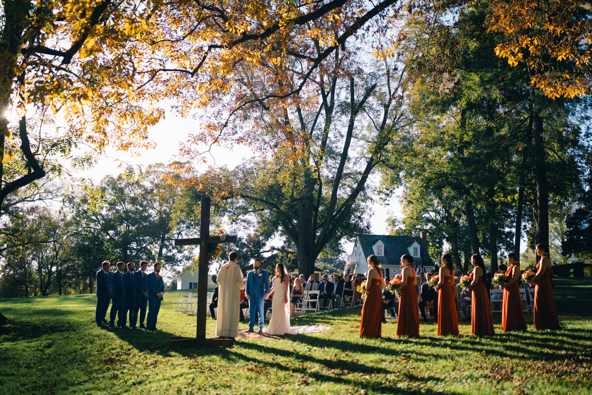 A rose hill estate wedding unfolds in a sunlit, outdoor setting with vibrant fall foliage. The couple stands by a wooden cross, surrounded by the bridal party in orange dresses and groomsmen in blue suits. Guests are seated on white chairs beneath the trees.