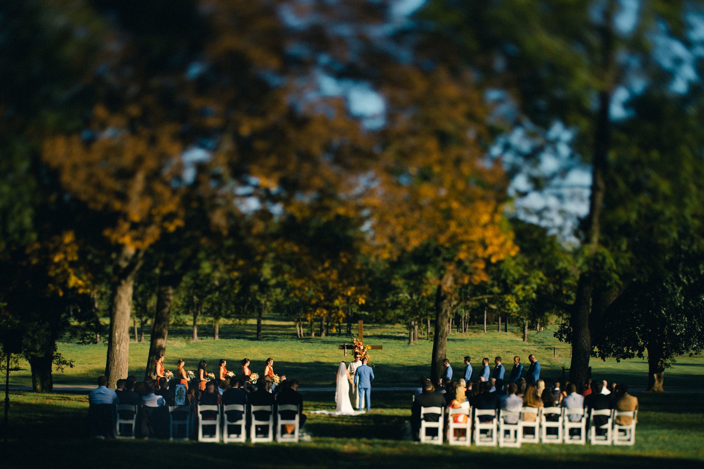 The wedding ceremony, set in a picturesque park reminiscent of a Rose Hill Estate wedding, features guests seated in rows flanking an aisle with tree-lined views. The couple stands at an altar adorned with flowers under the radiant, sunny sky.