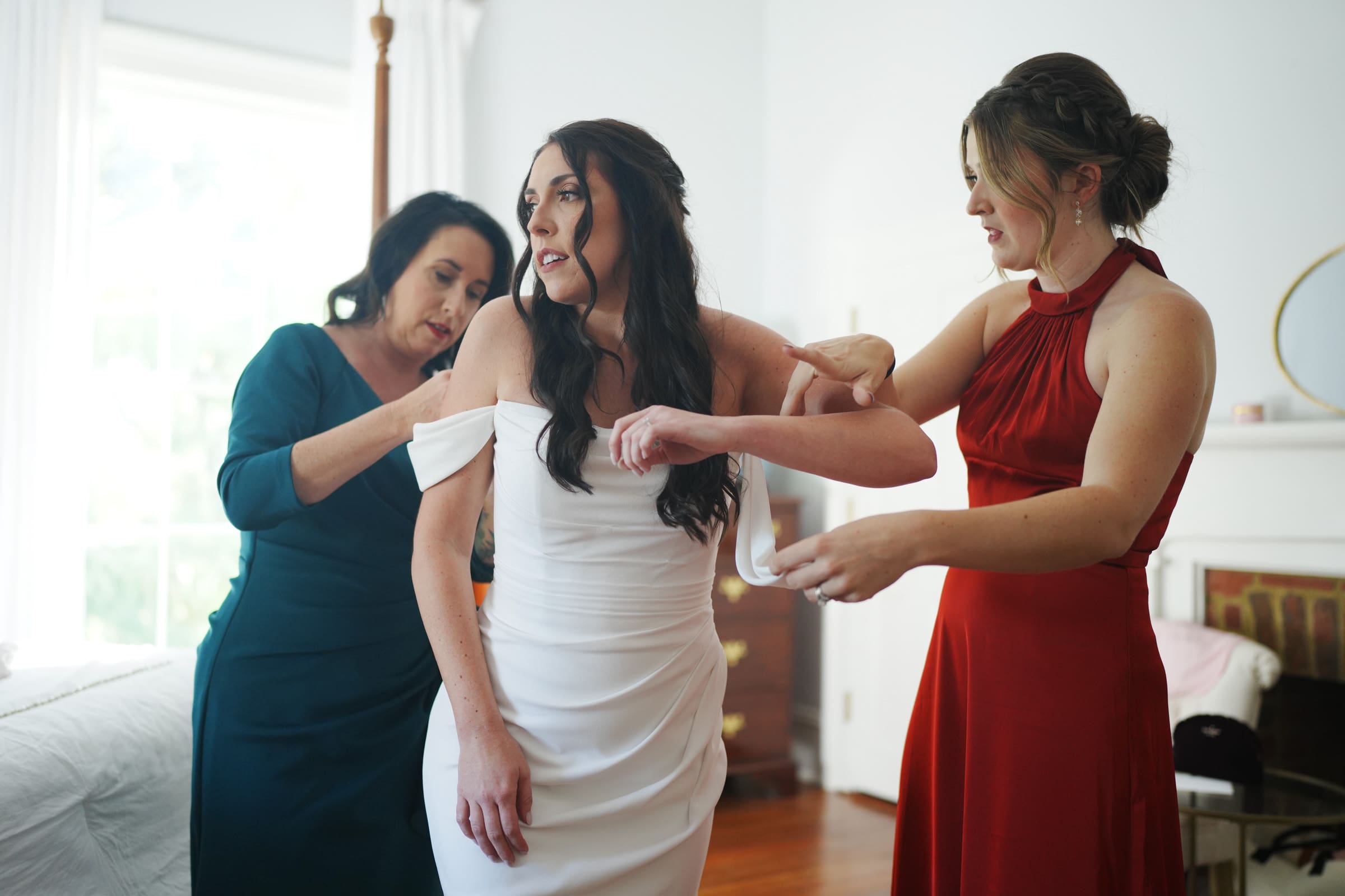 At a Rose Hill Estate wedding, the bride in a white dress is gracefully assisted by two women. One adjusts her sleeve, while the other secures a detail at the back. In the bright room, an atmosphere of focused support fills this enchanting moment.