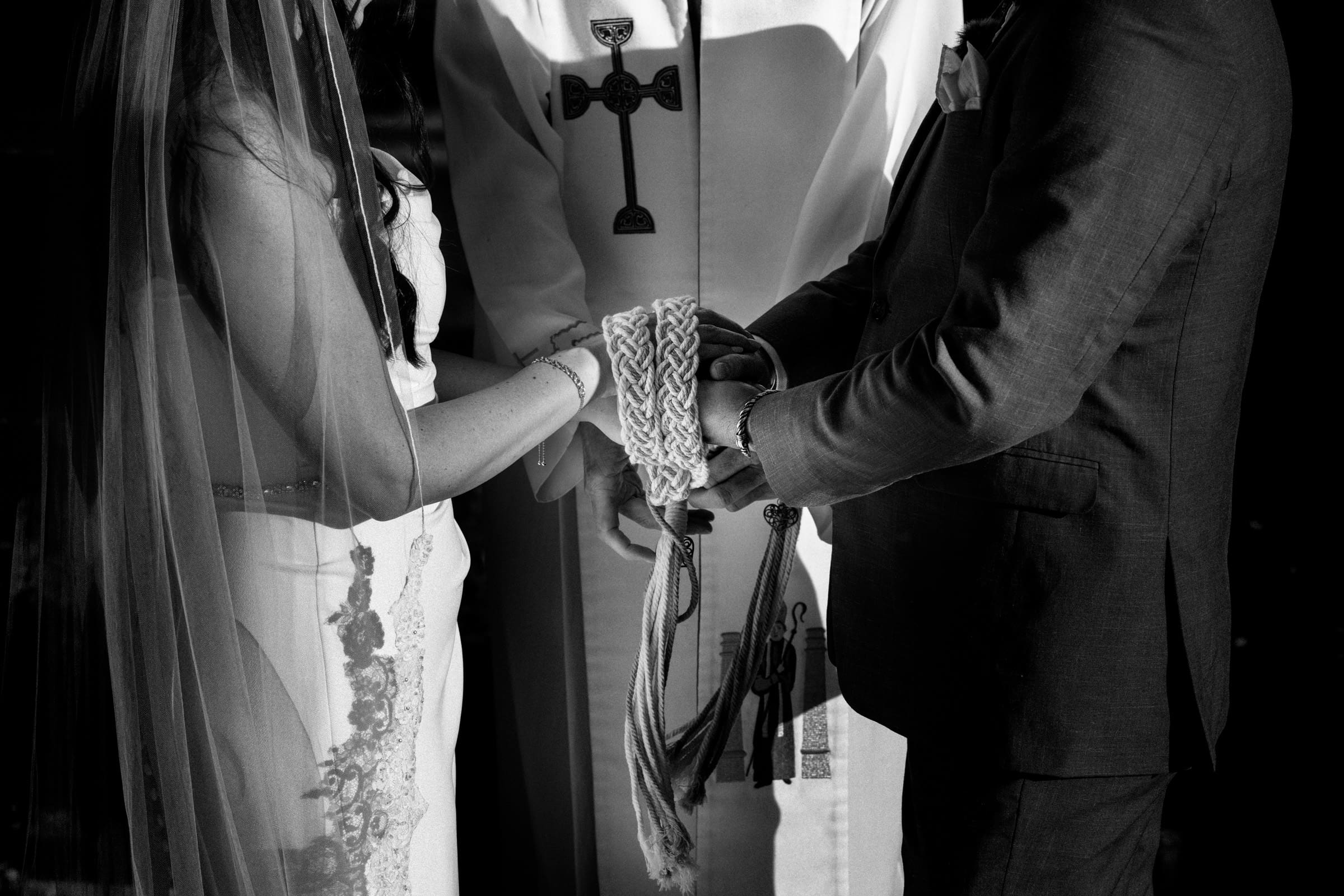 A couple is holding hands in a wedding ceremony at Rose Hill Estate, their hands wrapped in a braided cord. A person in religious attire stands behind them. The image is black and white, emphasizing the solemnity and significance of the moment.