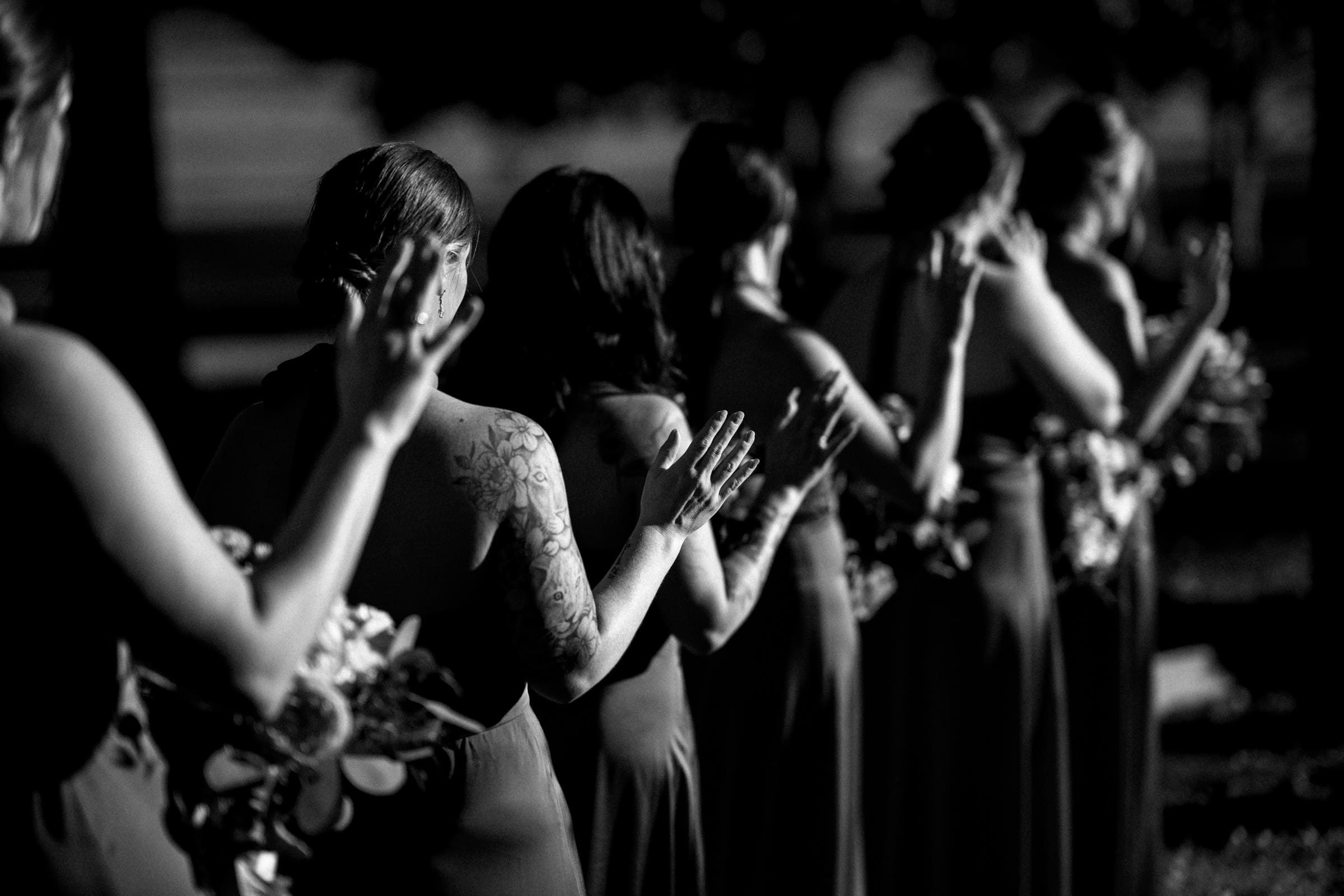A line of women in dresses seen from behind in black and white at a Rose Hill Estate wedding. They are holding bouquets and raising their hands, with the focus on the woman in the foreground who has a tattooed arm.