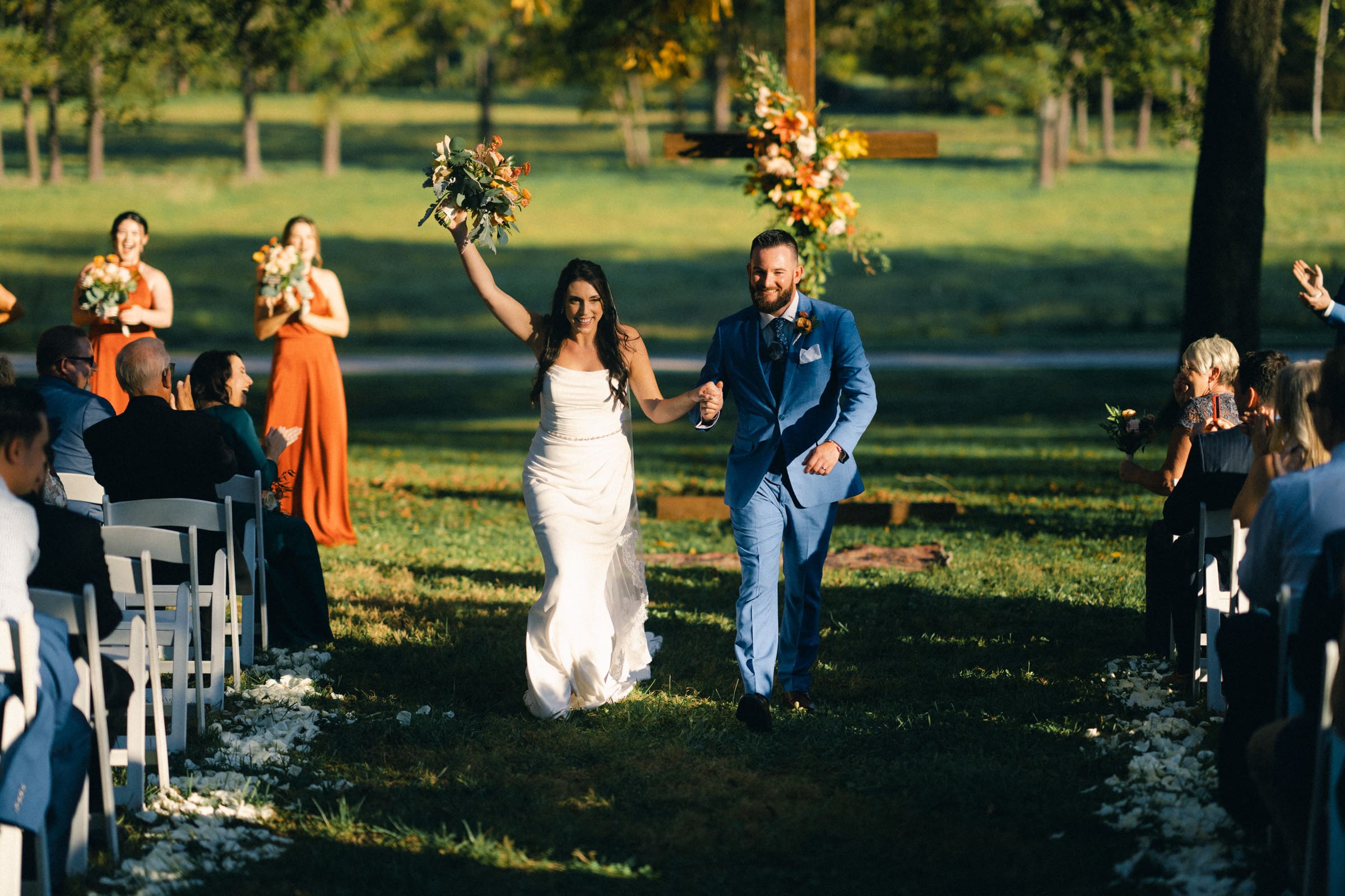 A joyful couple walks down the aisle at Rose Hill Estate, hand in hand. The bride in a white dress holds a bouquet overhead while the groom dons a blue suit. Guests seated on either side watch and celebrate amidst a backdrop of grassy fields and towering trees.