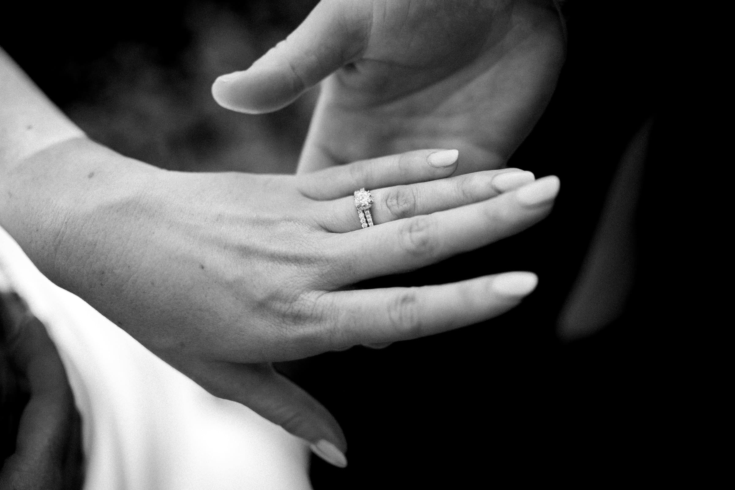 A black and white close-up image captures a couple's hands, one adorned with a diamond ring and elegantly manicured nails, the other gently supporting underneath. This intimate moment evokes the refined elegance of a Rose Hill Estate wedding, set against a dark background.