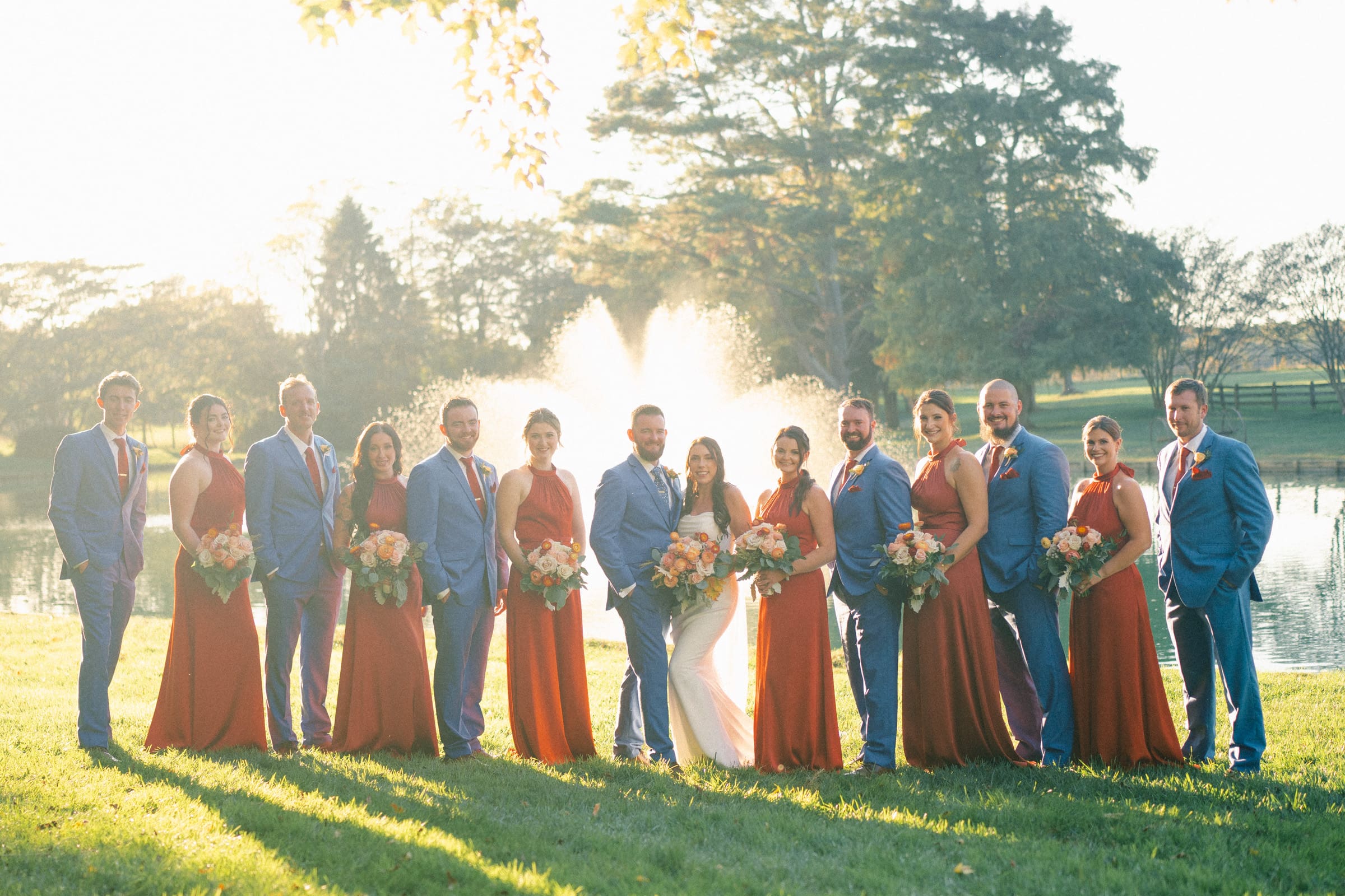 A wedding party poses outdoors at Rose Hill Estate in front of a fountain and trees. The bride is in a white dress, and the groom is in a blue suit. Bridesmaids in rust-colored dresses and groomsmen in blue suits with red ties stand on either side, all holding bouquets.