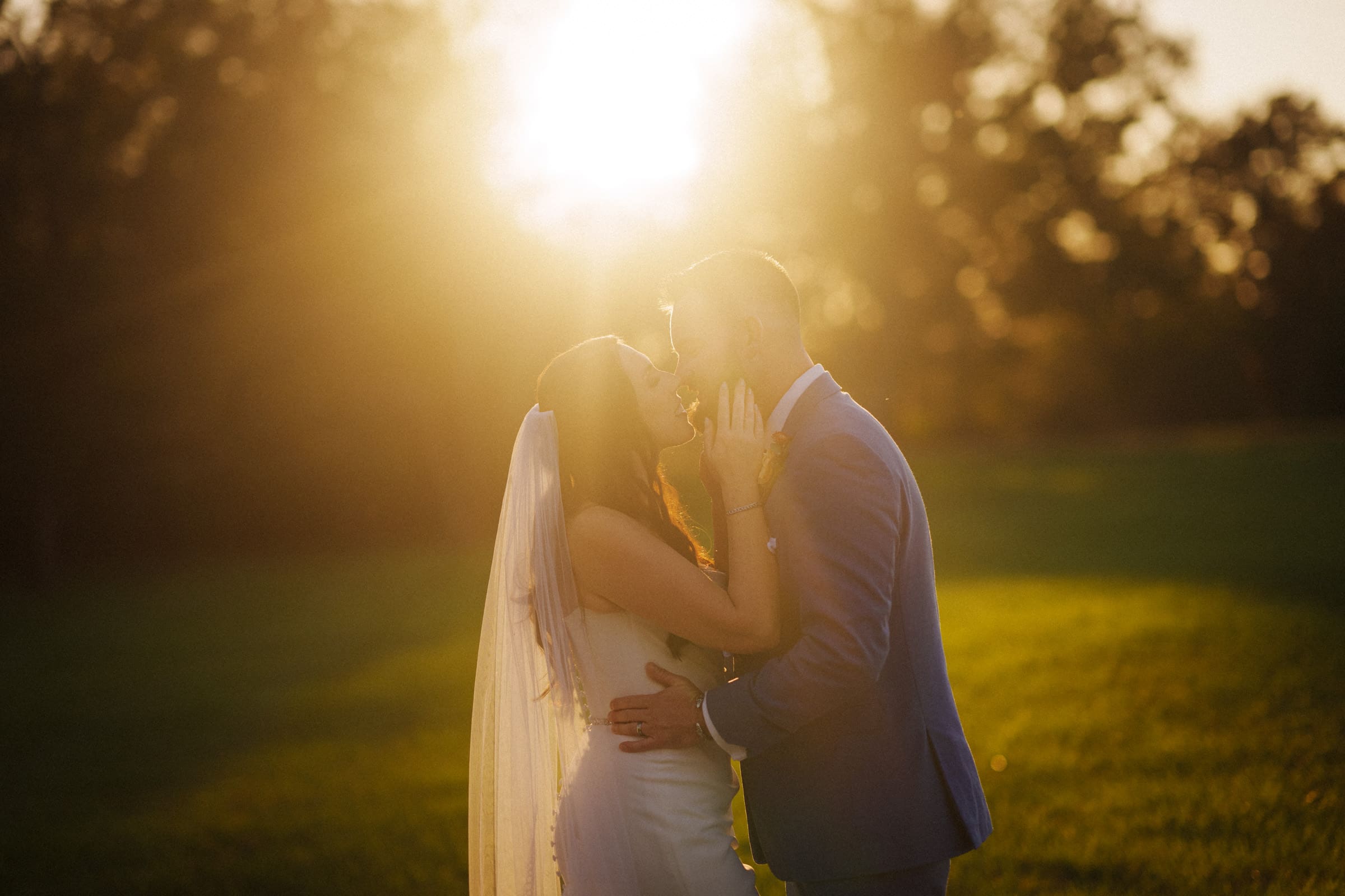 In a sunlit meadow at Rose Hill Estate, a bride and groom share a tender kiss. The bride's white dress and veil contrast beautifully with the groom's suit as the setting sun casts a warm, golden glow around them, casting long shadows on the grass.