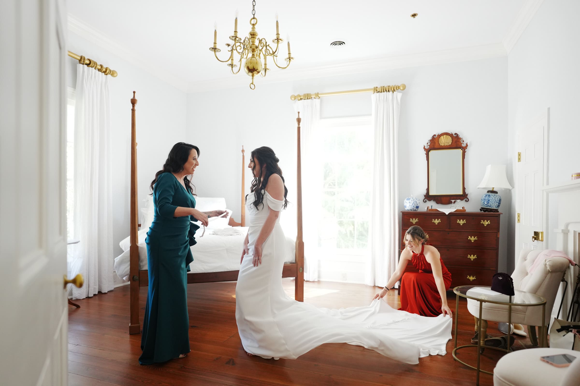 A bride in a white dress stands in the bright bedroom of Rose Hill Estate, surrounded by elegance. A woman in a green dress helps her, while another adjusts the train. The room features wooden floors, a chandelier, and a dresser with a mirror, capturing pure wedding day charm.