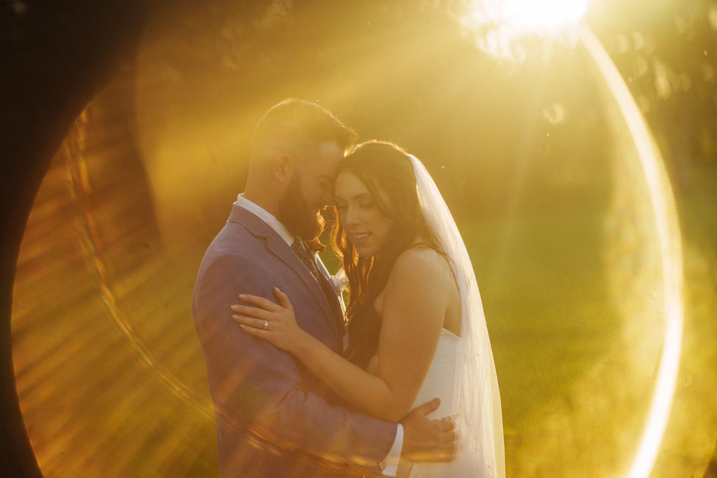 A bride and groom embrace tenderly in a sunlit field at Rose Hill Estate. The golden sunlight creates a warm, halo-like glow around them, enhancing the romantic atmosphere. The bride is wearing a veil, and the groom is in a suit, capturing the essence of an idyllic wedding day.