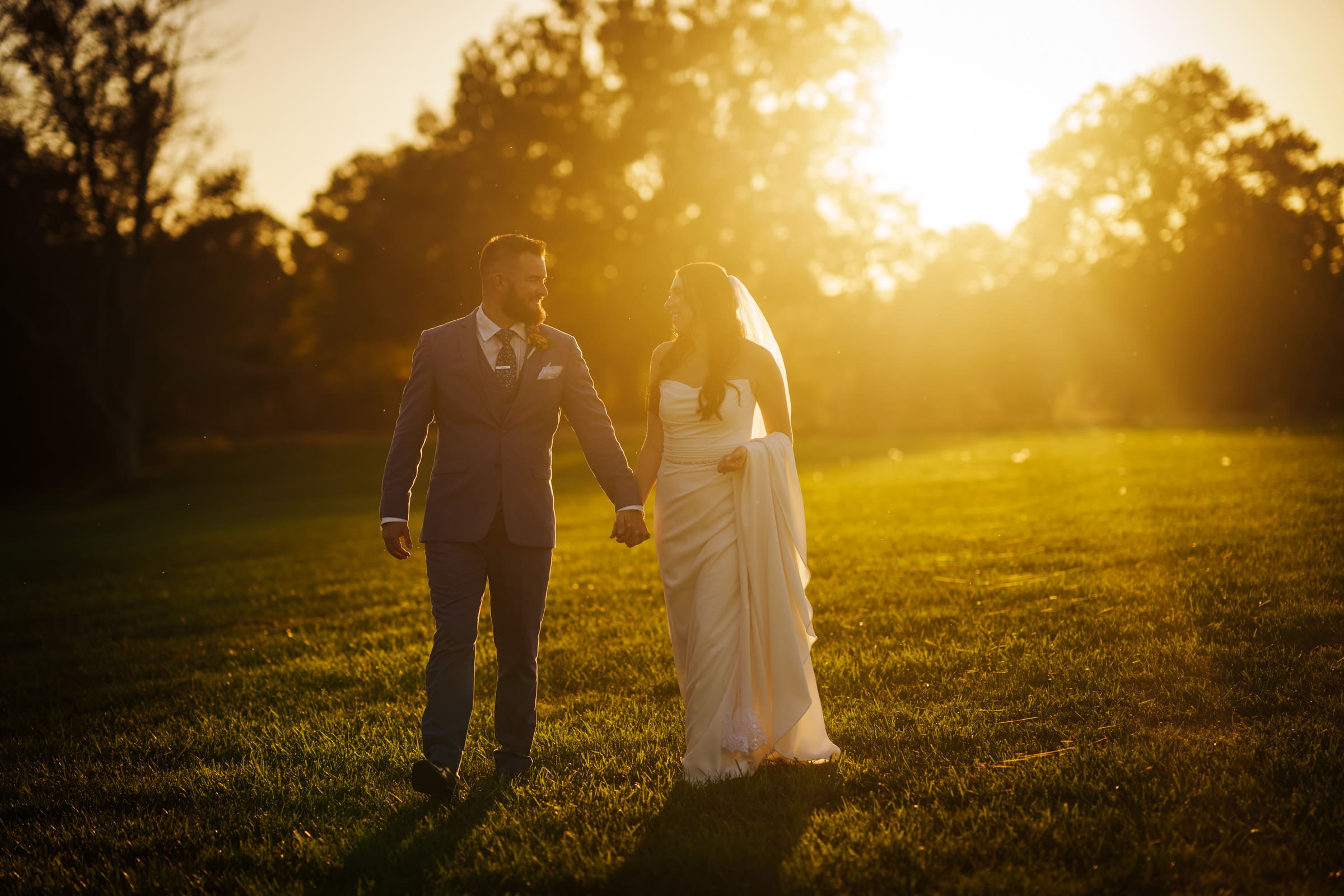 A couple in wedding attire strolls hand in hand across a sunlit grassy field at Rose Hill Estate during sunset. The groom is dapper in a gray suit, while the bride dazzles in her white dress and veil. Trees and a golden sky create a picturesque backdrop.