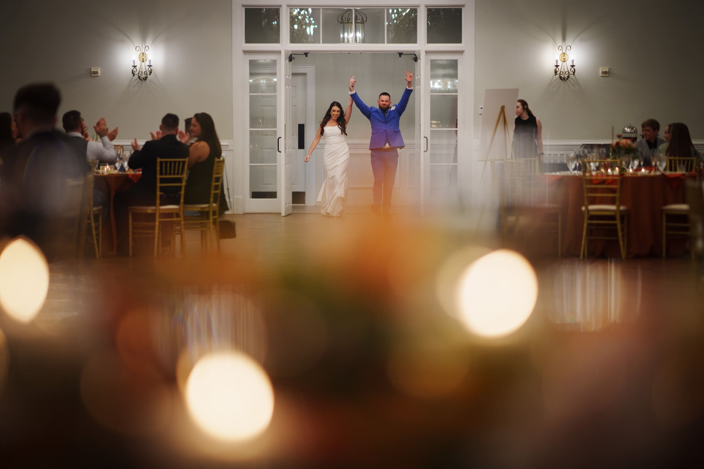 At the Rose Hill Estate wedding, a bride in a white dress and a groom in a blue suit joyfully enter the warmly lit banquet hall, greeted by seated guests. Soft, out-of-focus lights frame the foreground, adding to the festive atmosphere.