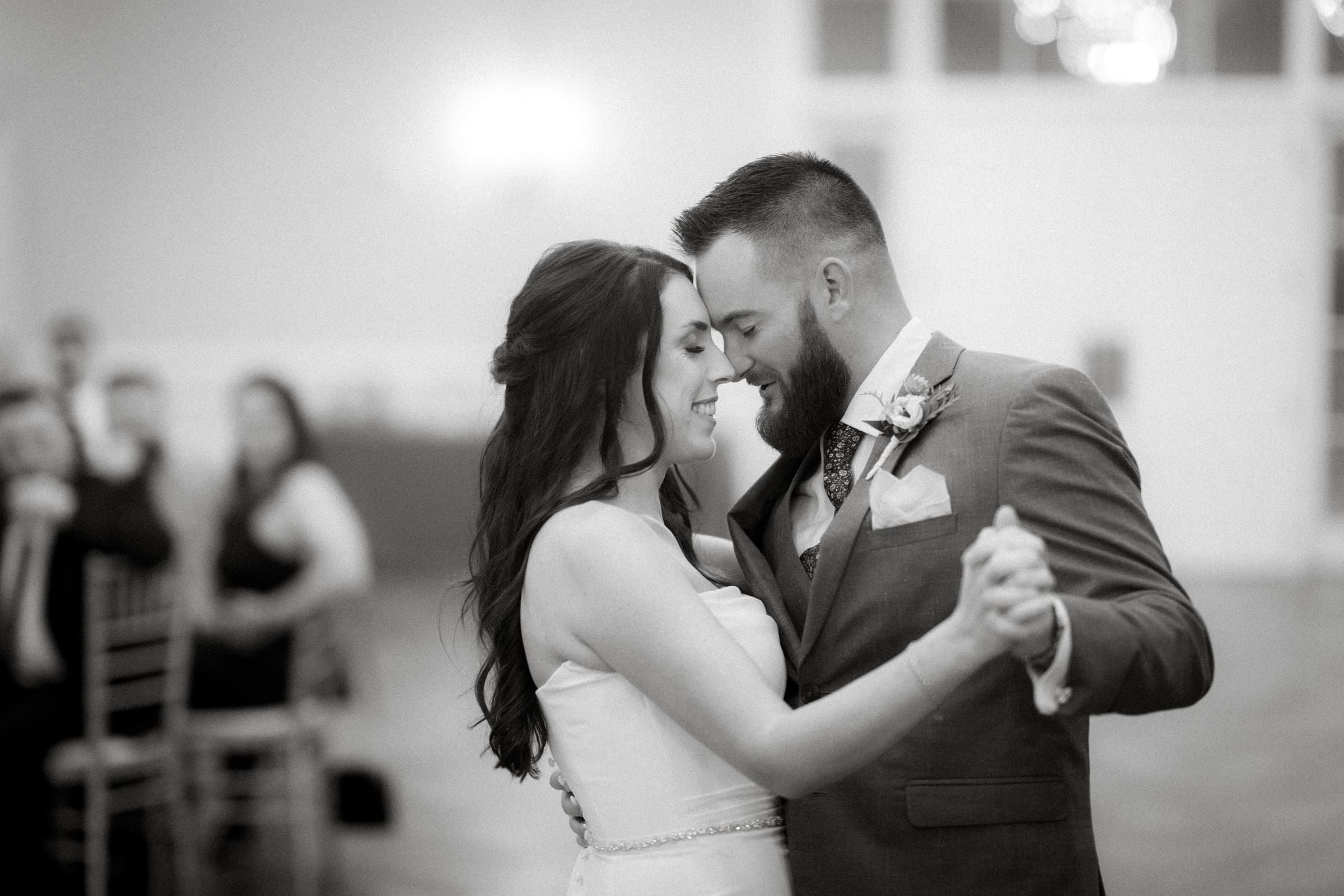 At their Rose Hill Estate wedding, the bride and groom share a joyful first dance. The bride wears a strapless dress while the groom dons a suit with a boutonniere. Guests are seated, enjoying the softly lit ambiance in the background.