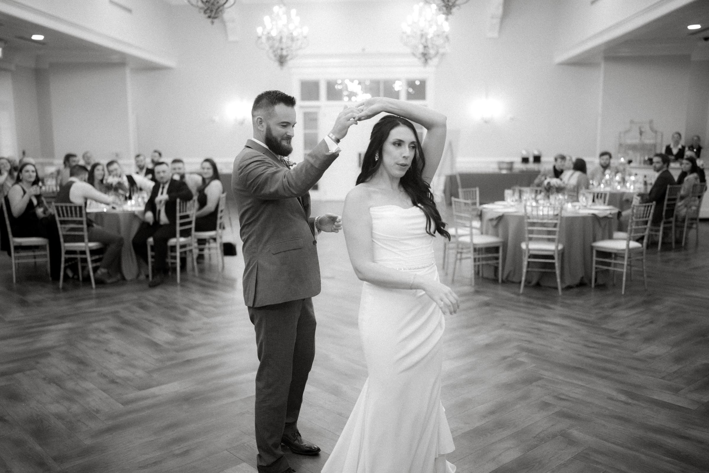 In the grand elegance of Rose Hill Estate, a couple dances gracefully on a wooden floor. The man twirls the woman in her long dress, captivating seated guests who watch intently. Chandeliers and round tables add to the enchanting wedding scene.