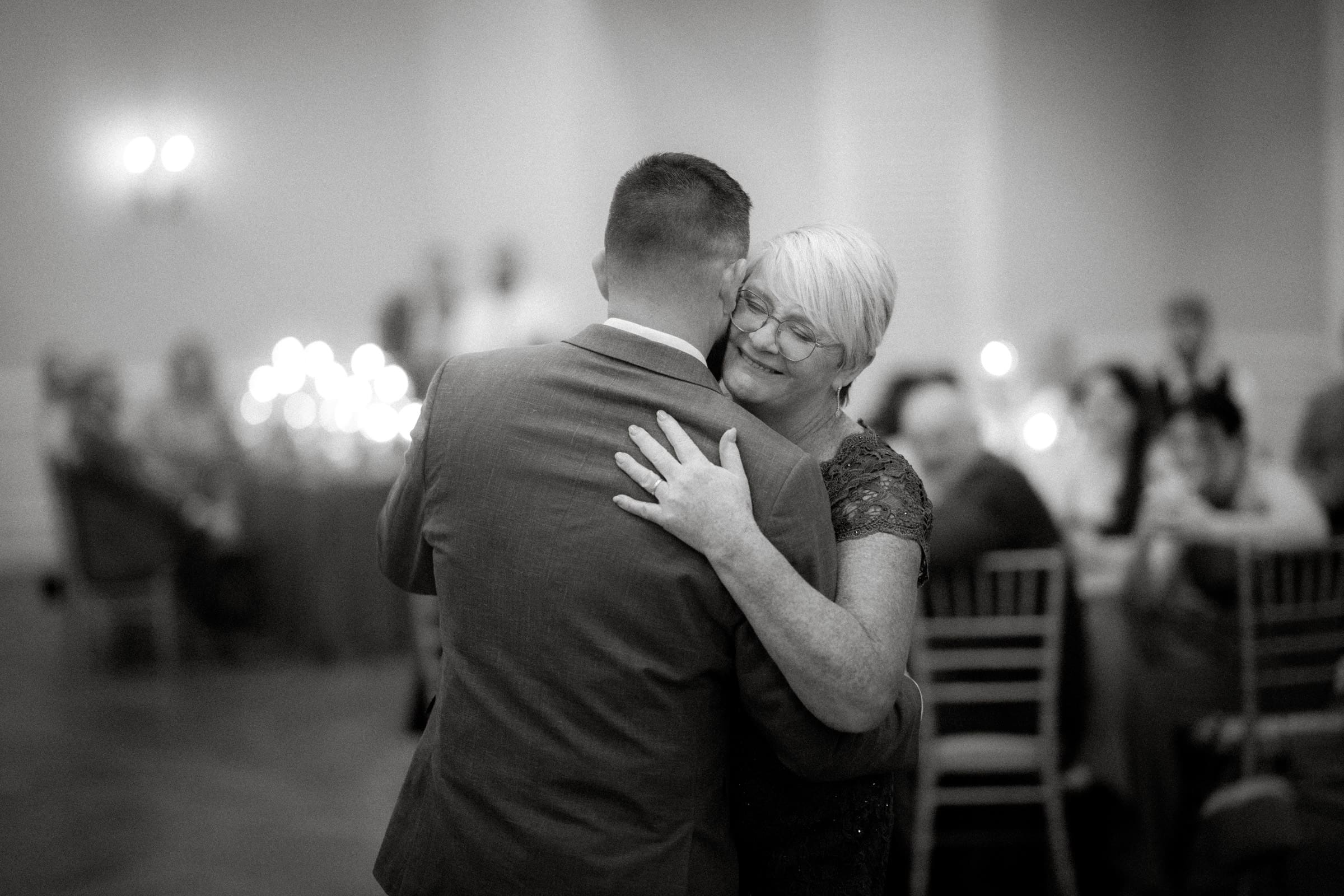 A black-and-white photo captures an elderly woman and a man dancing closely at a formal Rose Hill Estate wedding. She has a gentle smile and her eyes closed, while in the background, guests are seated at tables with candles softly glowing.