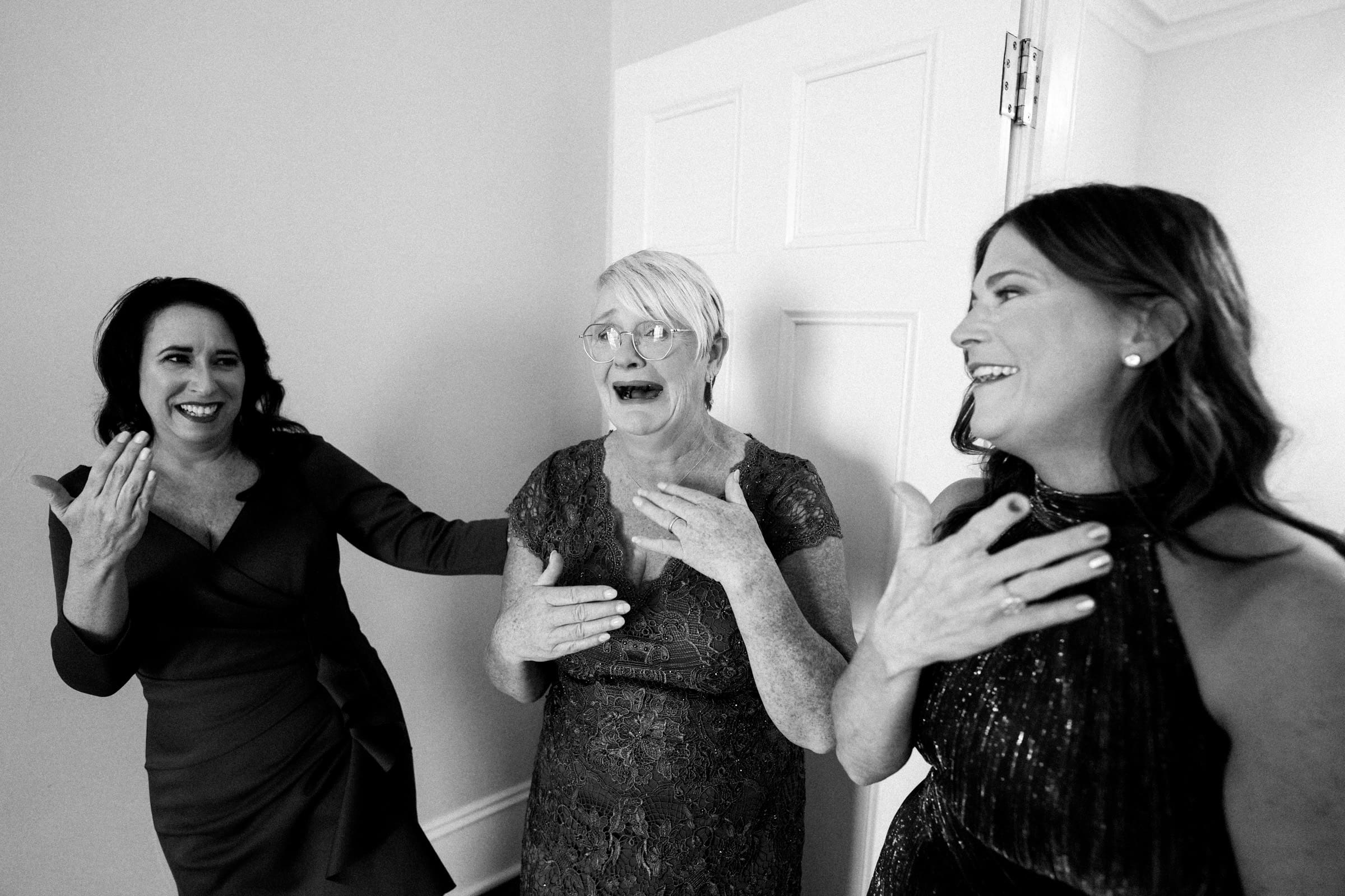 In the elegant setting of Rose Hill Estate, three women stand together, smiling and laughing. One has her hand on her chest, the older woman in the middle exudes joy, and the third covers her mouth in delight, all adorned in formal attire perfect for a wedding.