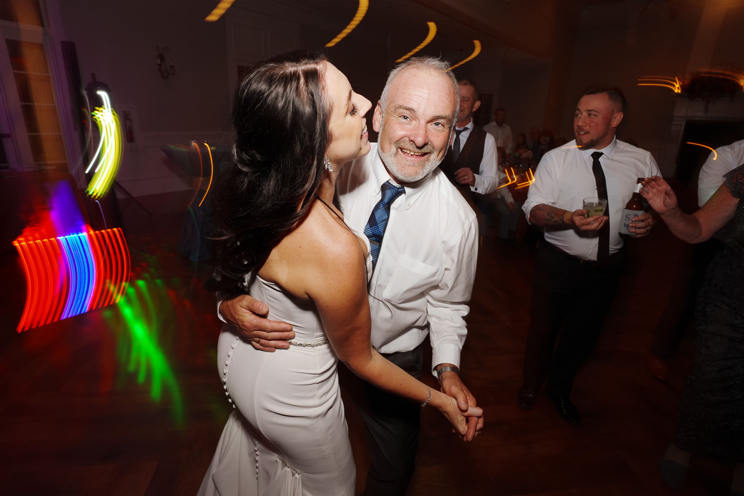At the Rose Hill Estate wedding, a woman in a white dress smiles and leans in to kiss the cheek of a man in a white shirt and tie as they dance in the dimly lit room. Others are dancing in the background, colorful light trails adding to the lively atmosphere.