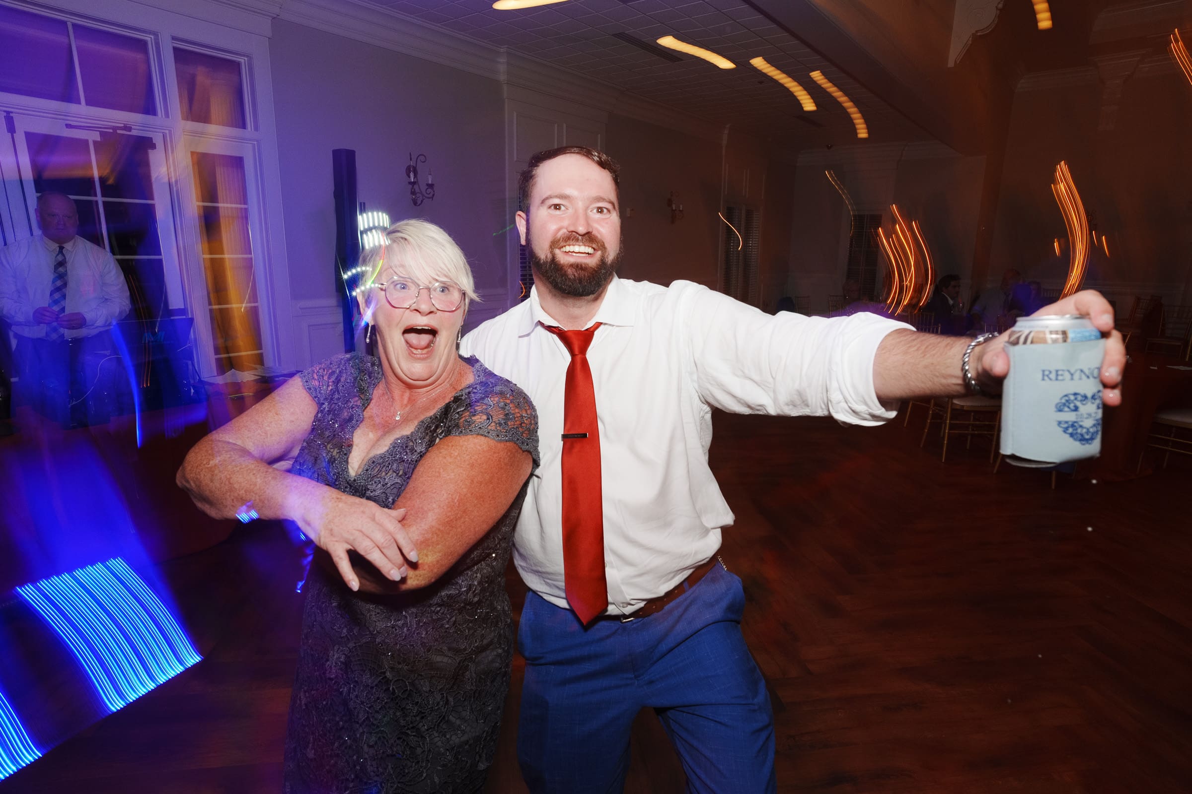 A woman in a purple dress and a man with a red tie and white shirt pose joyfully on the dance floor at the Rose Hill Estate wedding. The man holds a drink can with REYN partially visible in the dimly lit room, where bright lights create an enchanting motion blur.