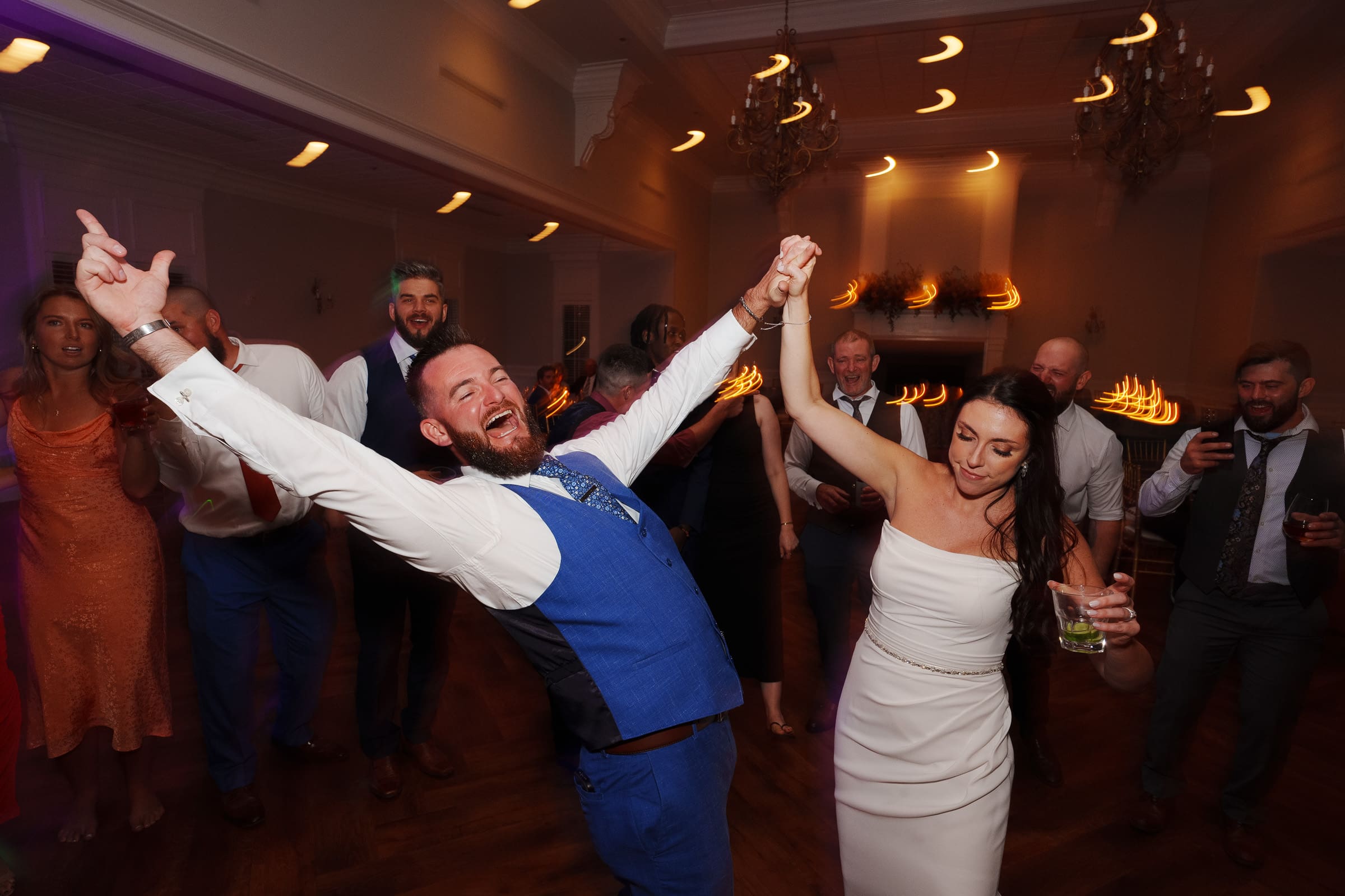 At their Rose Hill Estate wedding, the groom in a blue suit and bride in a white dress dance joyfully, holding hands and raising their arms. Surrounded by smiling guests on the warmly lit dance floor, some guests clap beneath the chandeliers.
