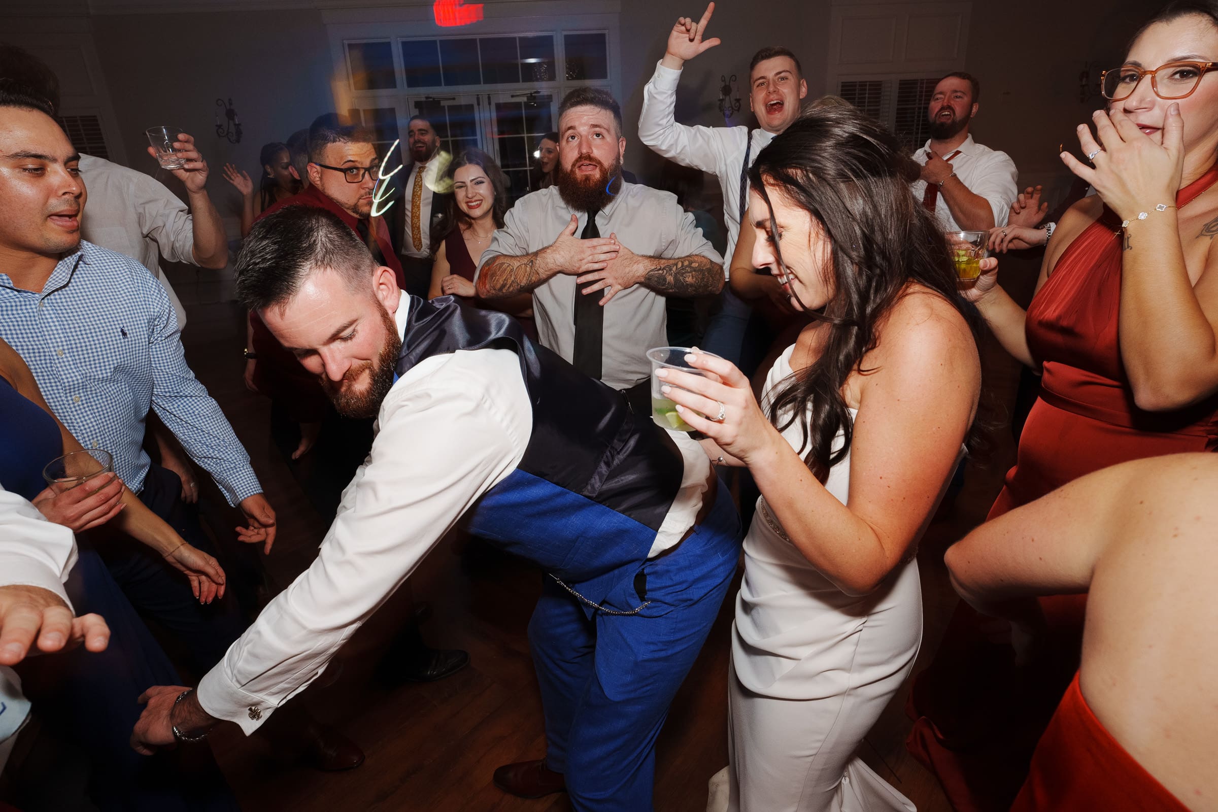 A lively Rose Hill Estate wedding reception has guests dancing energetically. A man in a suit leans forward playfully while others around him laugh and enjoy drinks. The dimly lit room exudes a joyful and celebratory atmosphere, perfect for this special occasion.