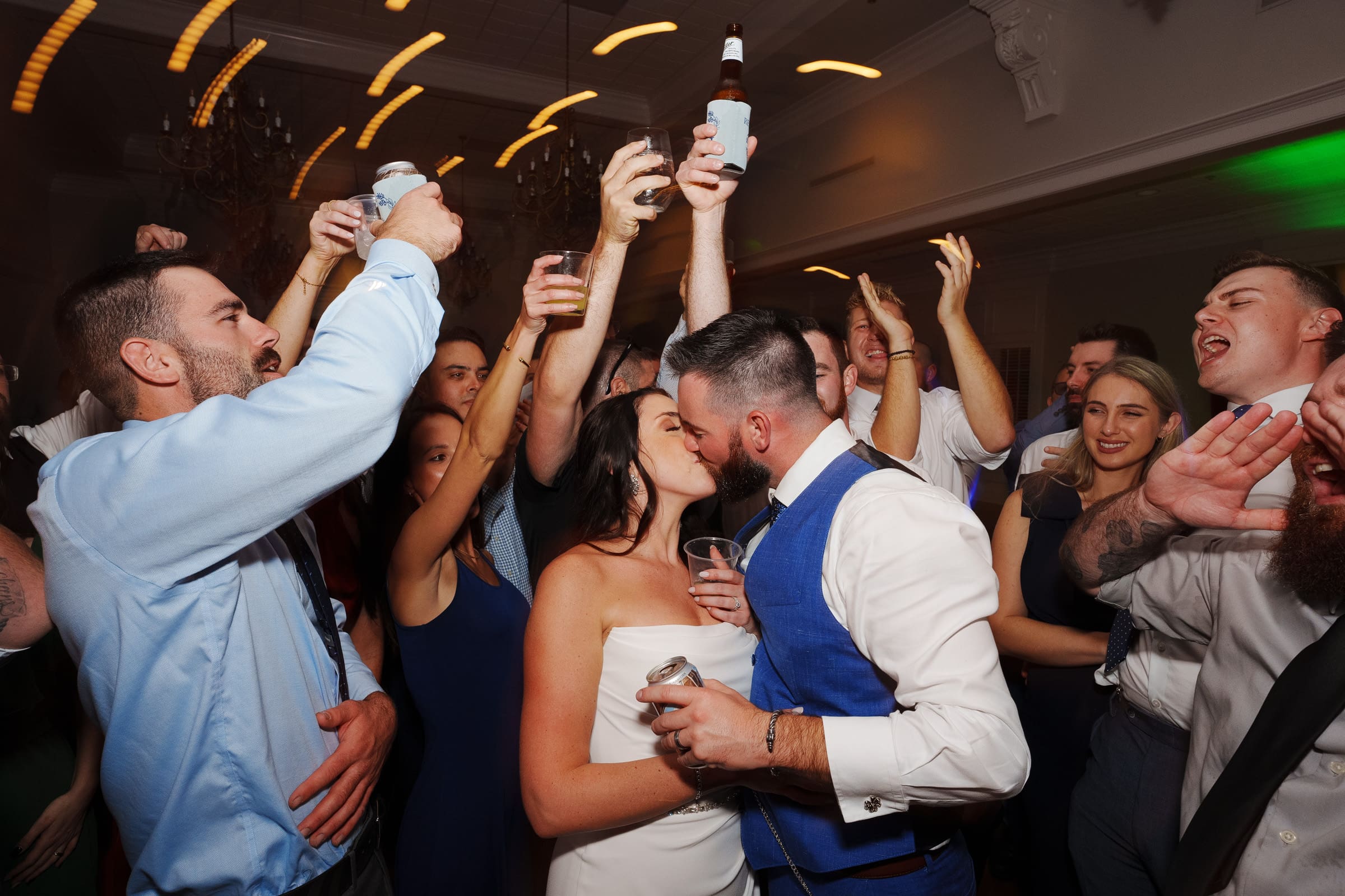 A newlywed couple shares a kiss on the dance floor at their picturesque Rose Hill Estate wedding, surrounded by friends and family celebrating. Guests raise drinks in the air, smiling and embracing the lively atmosphere of the unforgettable reception.
