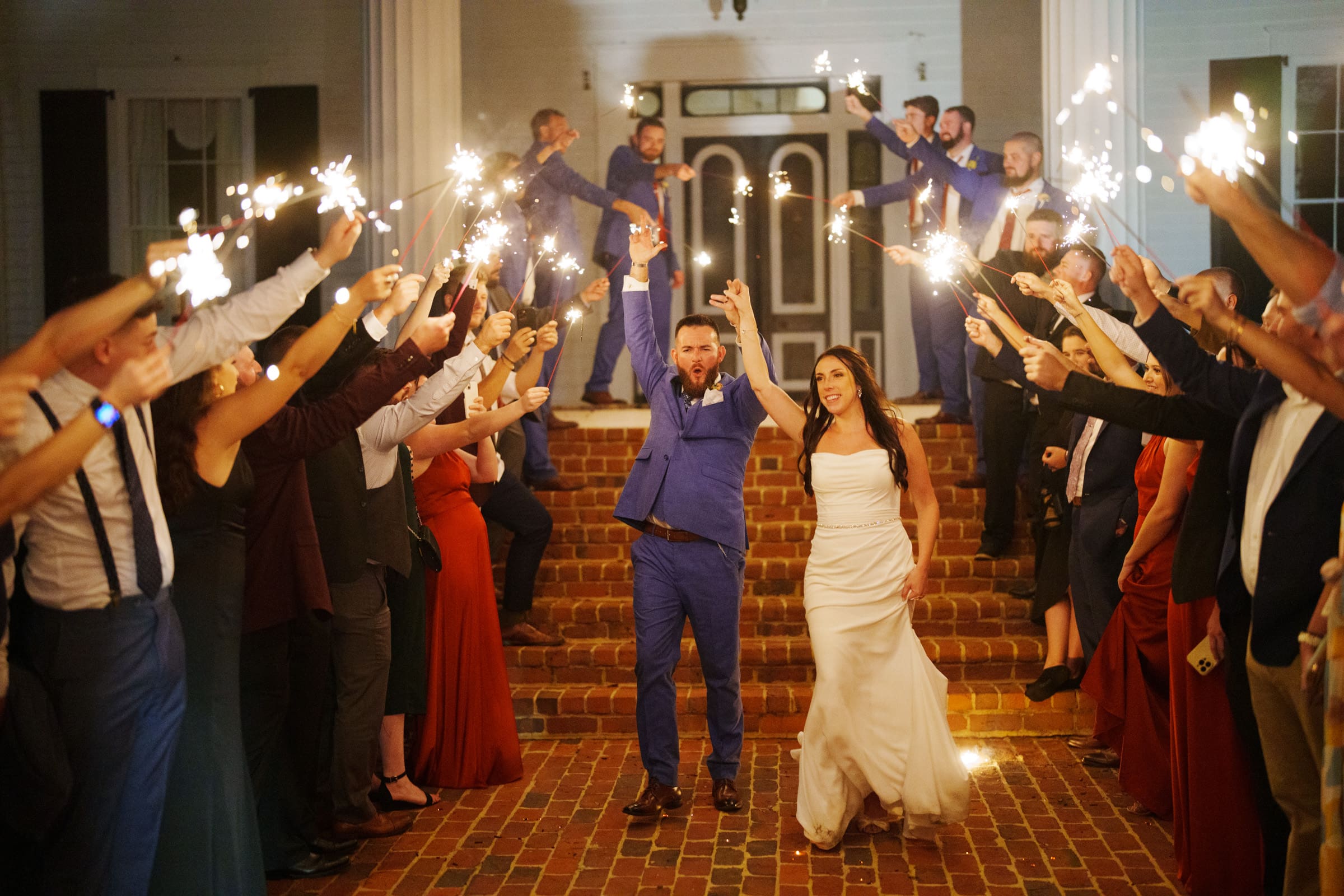 At their enchanting Rose Hill Estate wedding, a bride and groom joyfully walk through a sparkler-lit path created by cheering guests on a brick walkway. The groom is wearing a blue suit, and the bride is dressed in a white gown as guests line both sides, holding sparklers.