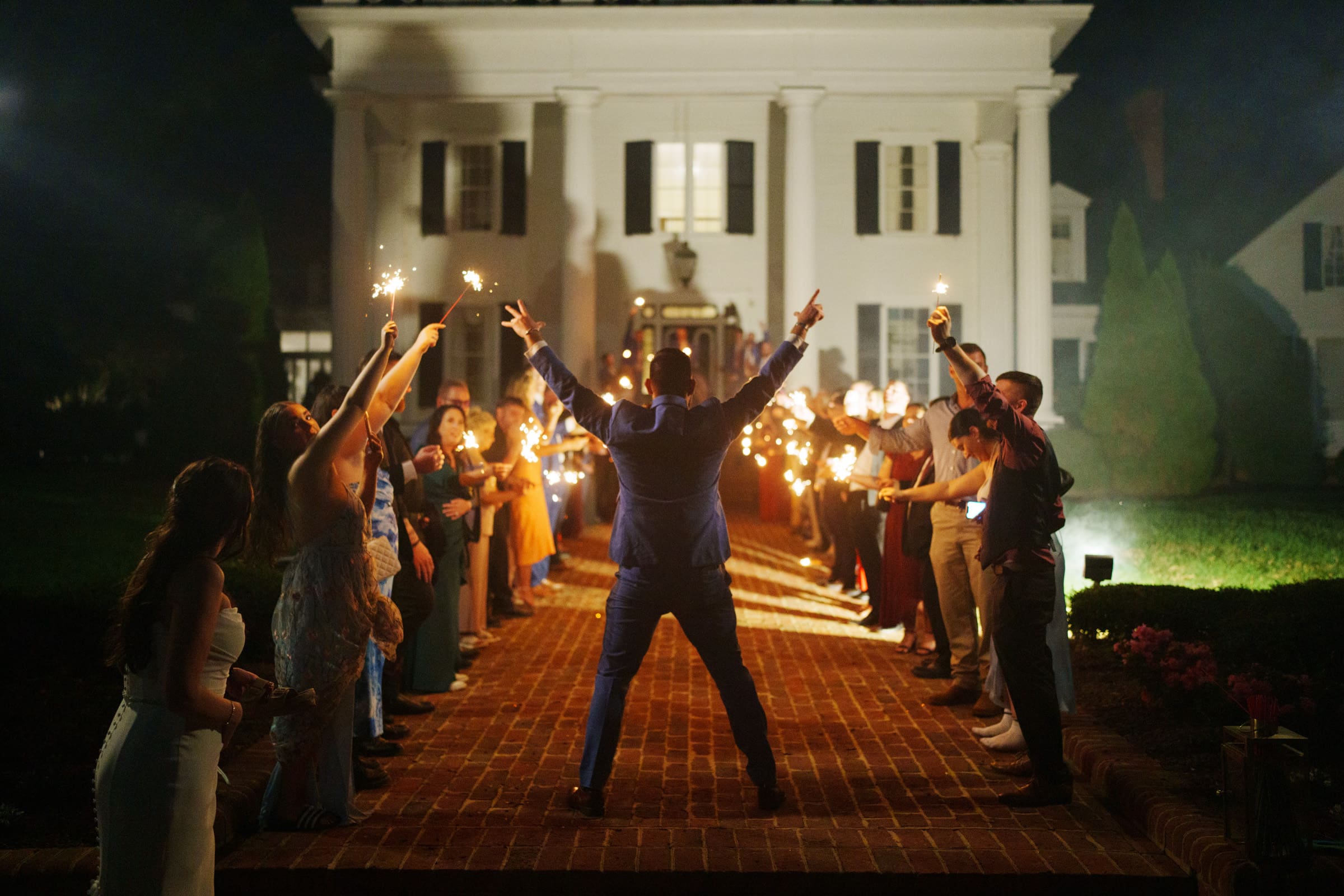 A person in a blue suit stands with arms raised in celebration at a Rose Hill Estate wedding, surrounded by a jubilant group holding sparklers. They are on a brick path leading to the beautifully illuminated building at night.