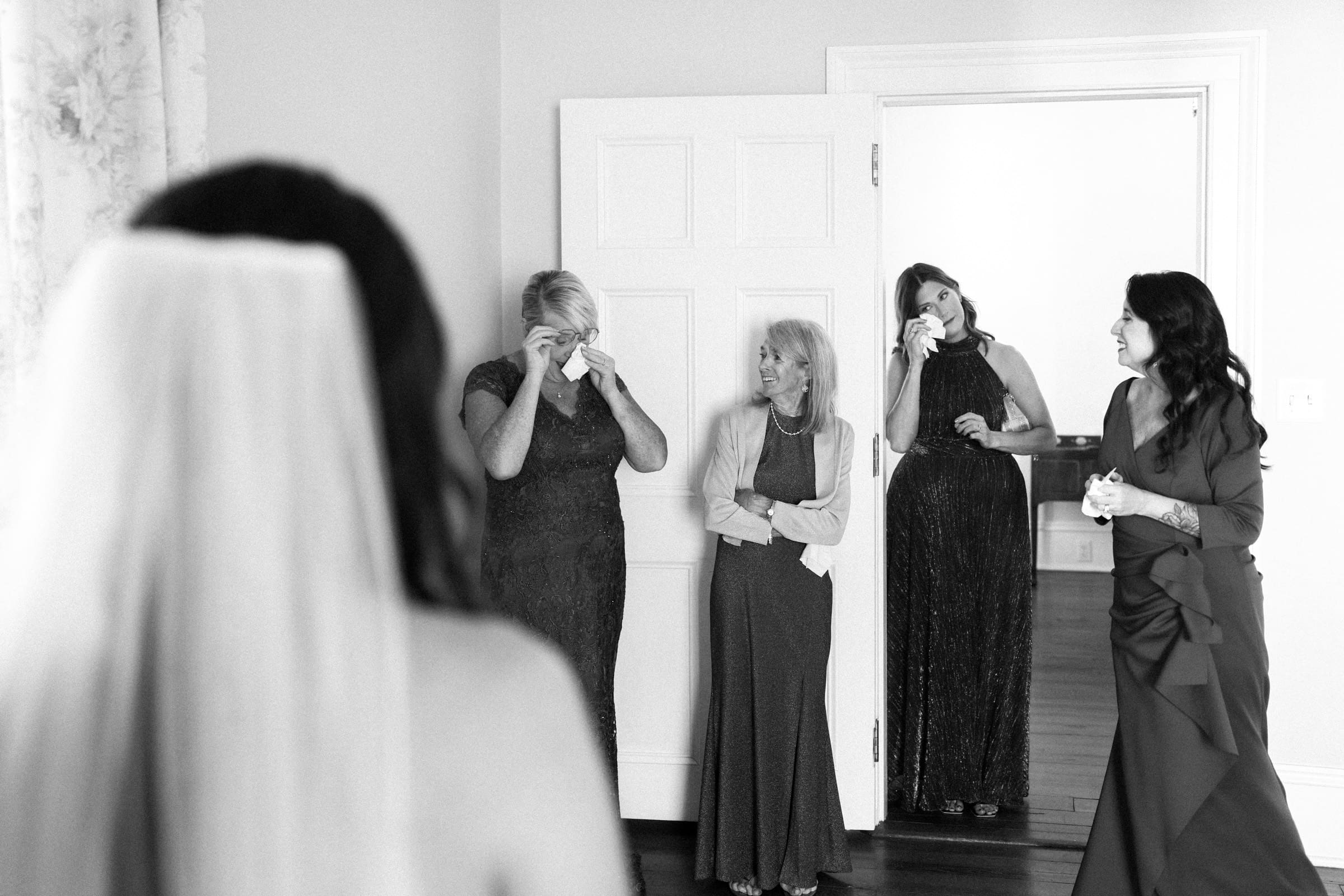 A black-and-white photo captures a poignant moment at a Rose Hill Estate wedding. The bride stands with her back to the camera as four women gather near a door, two wiping tears and one elderly woman smiling, all visibly moved at the sight of the bride in her dress.