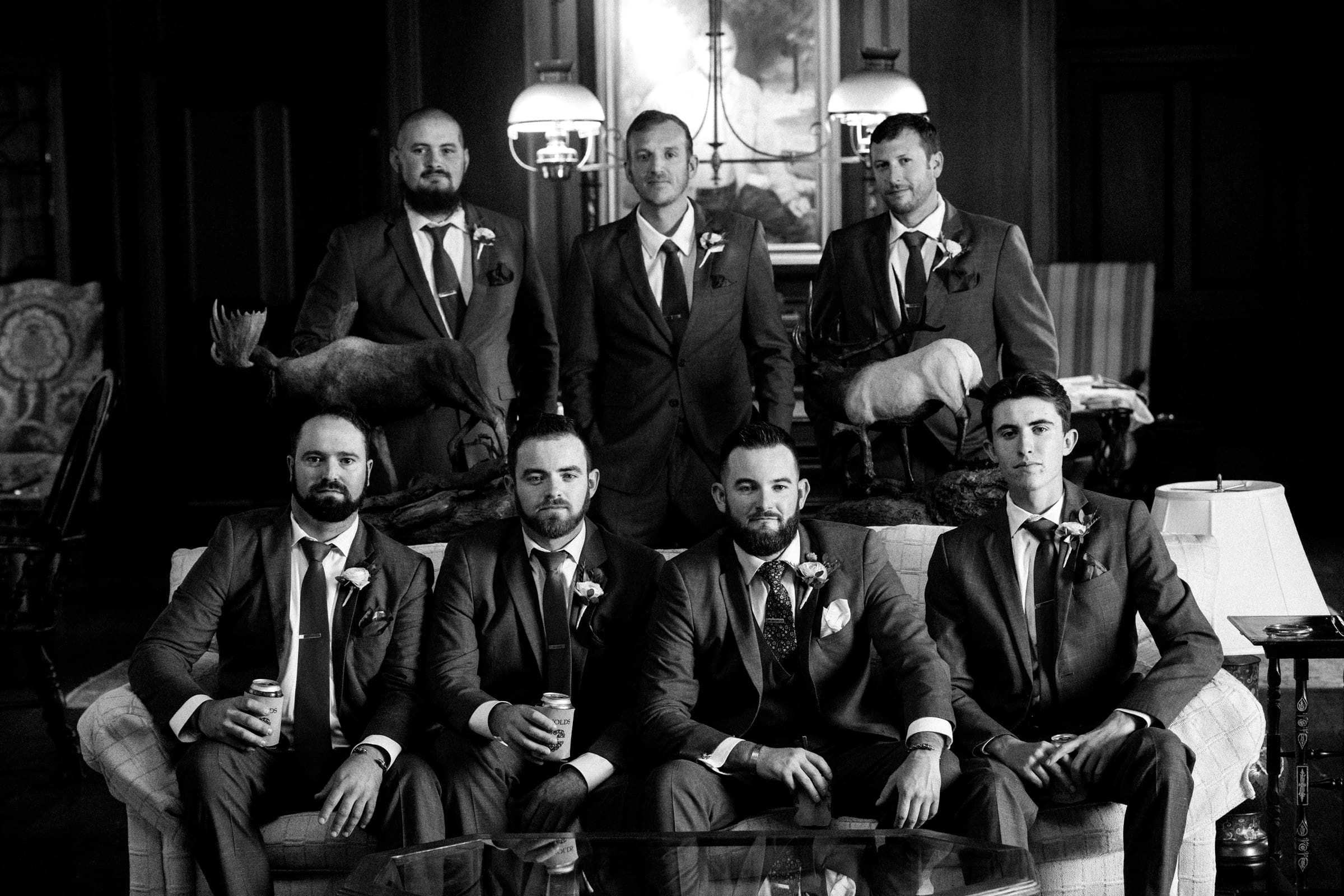 A black and white photo captures seven men in suits, adorned with boutonnieres, gathered around a sofa in a formal room at Rose Hill Estate. The mounted animal heads in the background add an elegant touch to this wedding scene.