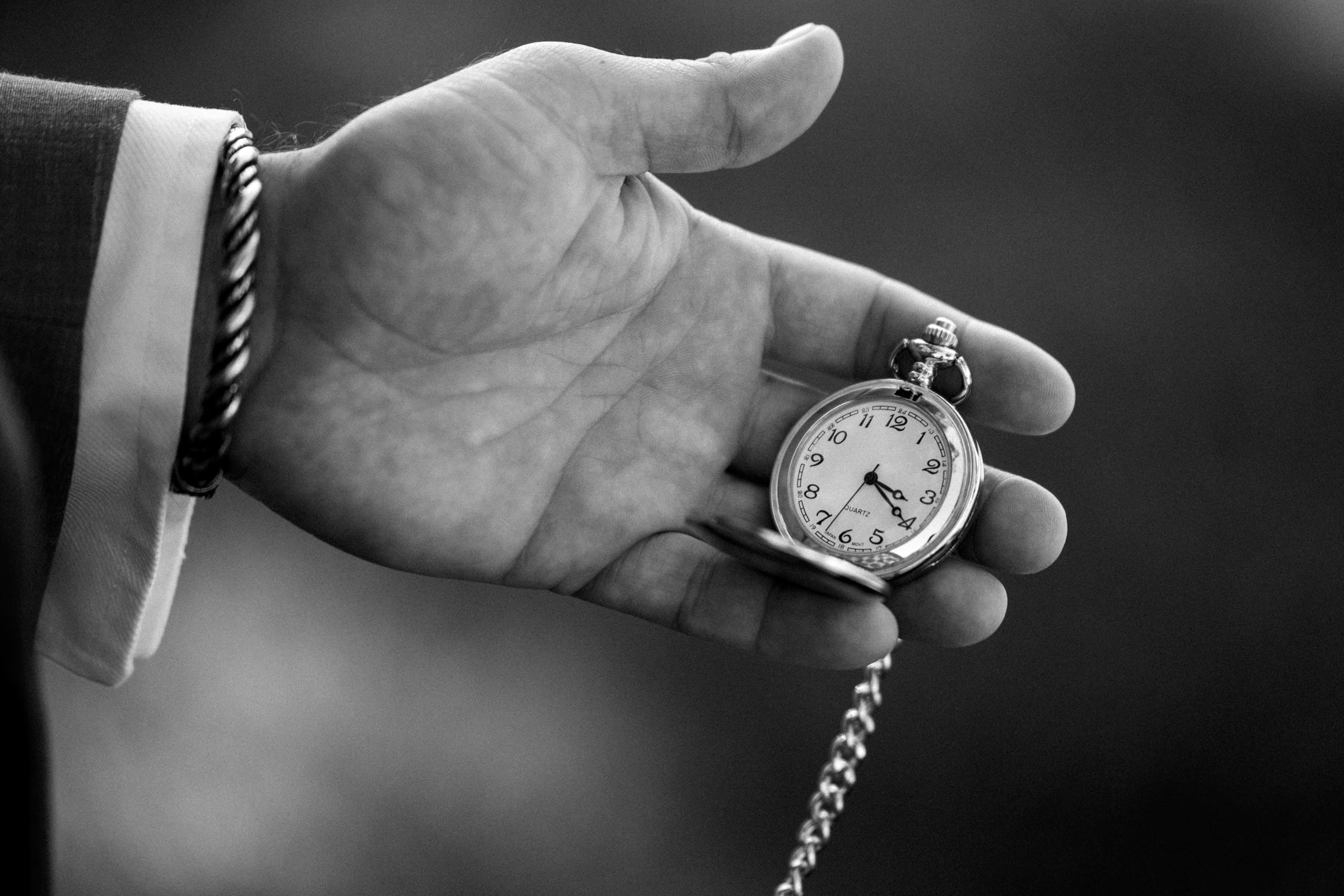 A black and white image captures a hand holding an open pocket watch, its Roman numerals and ornate hands glinting softly, reminiscent of a timeless Rose Hill Estate wedding. The watch is attached to a chain, the person’s suit sleeve visible beneath the elegant cuff.
