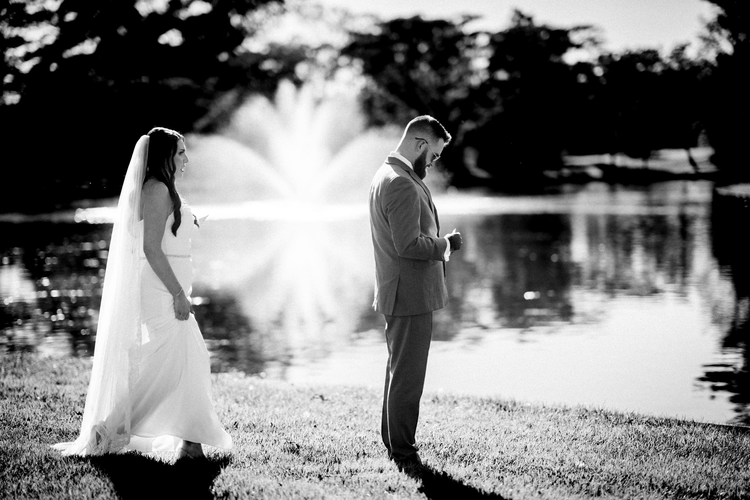 A bride and groom stand in a garden by a lake with a fountain at Rose Hill Estate, surrounded by trees. The groom, in a suit, looks down while the bride, in a dress and veil, stands behind him. It's a sunny day at the estate, creating a serene and romantic atmosphere.