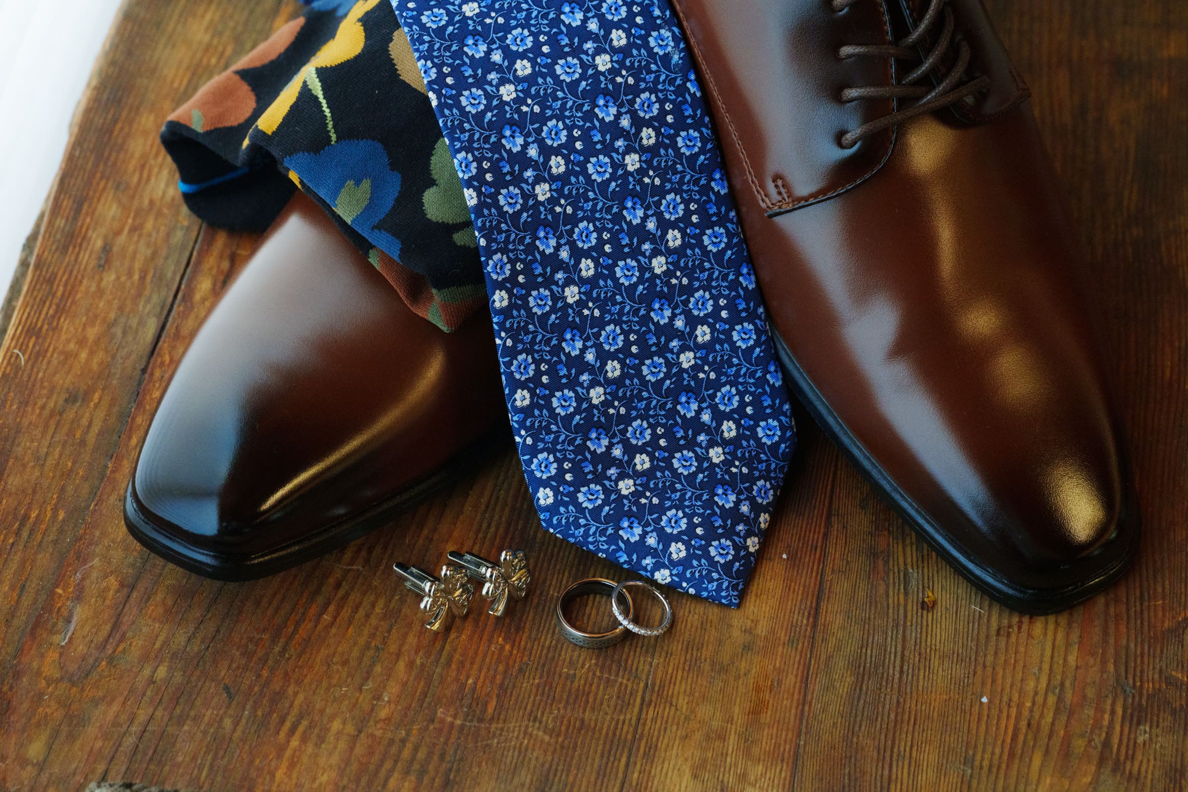 Brown leather shoes, a blue floral tie, a multicolored pocket square, two rings, and cufflinks are elegantly arranged on a wooden surface, capturing the charm of a Rose Hill Estate wedding.