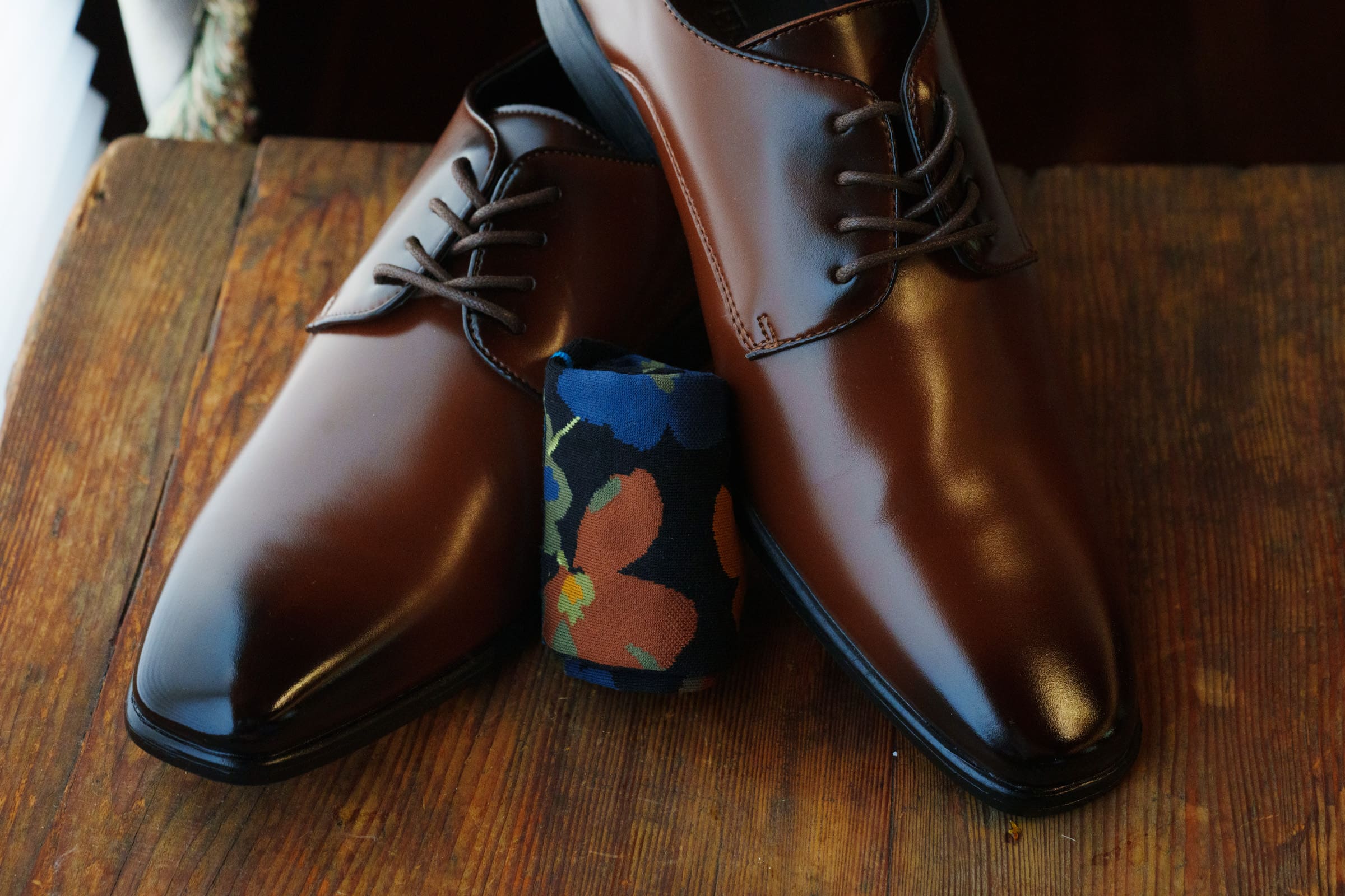 A pair of shiny brown leather dress shoes with laces rests on a wooden surface, perfect for a Rose Hill Estate wedding. Between the shoes lies a rolled-up sock featuring a vibrant floral pattern in blue, orange, and green.
