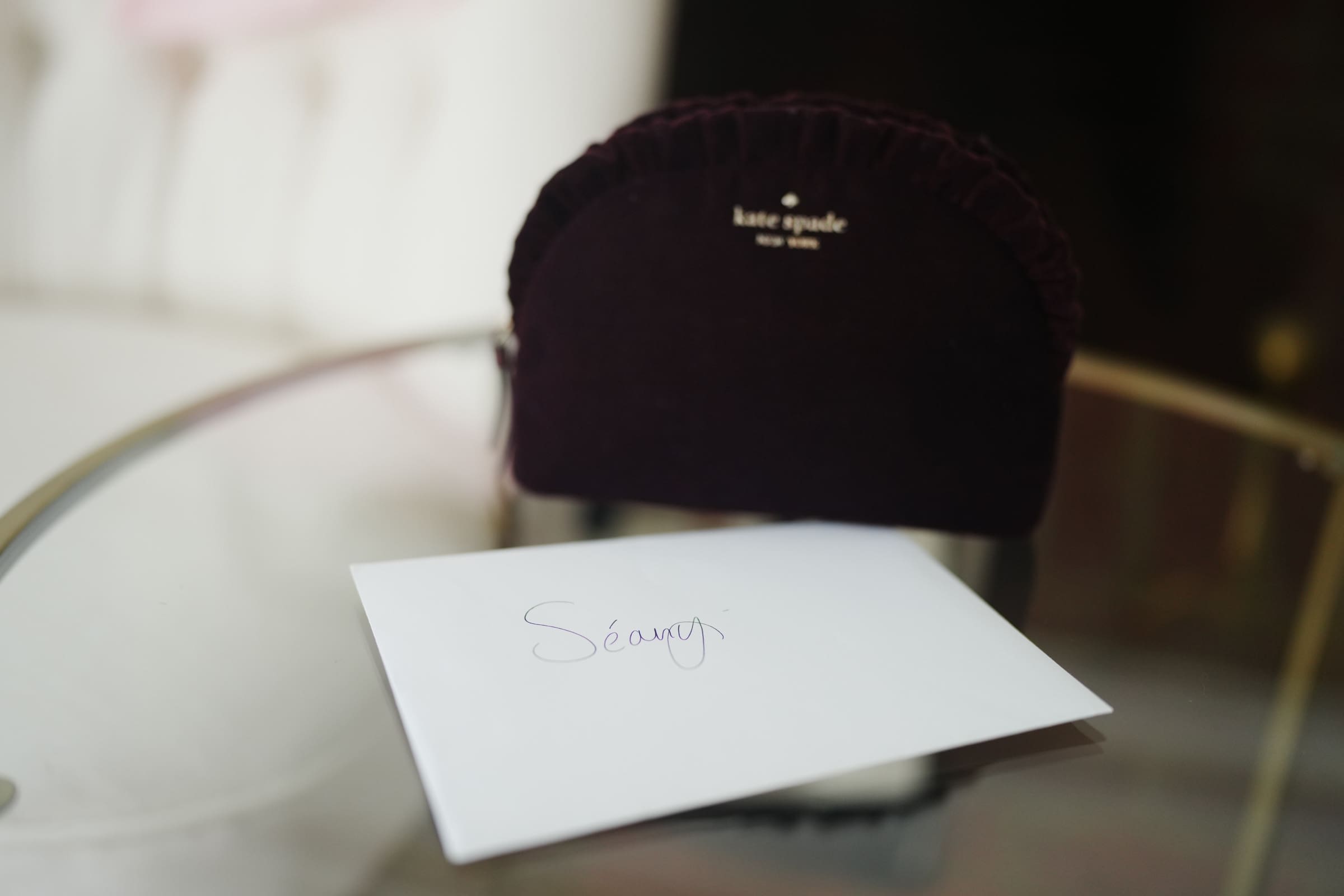 A small black Kate Spade pouch sits on a glass table, reminiscent of the elegance found at a Rose Hill Estate wedding. In front of it is a white envelope with the handwritten name Séamus on it. The background is blurred, adding an air of mystery and romance.