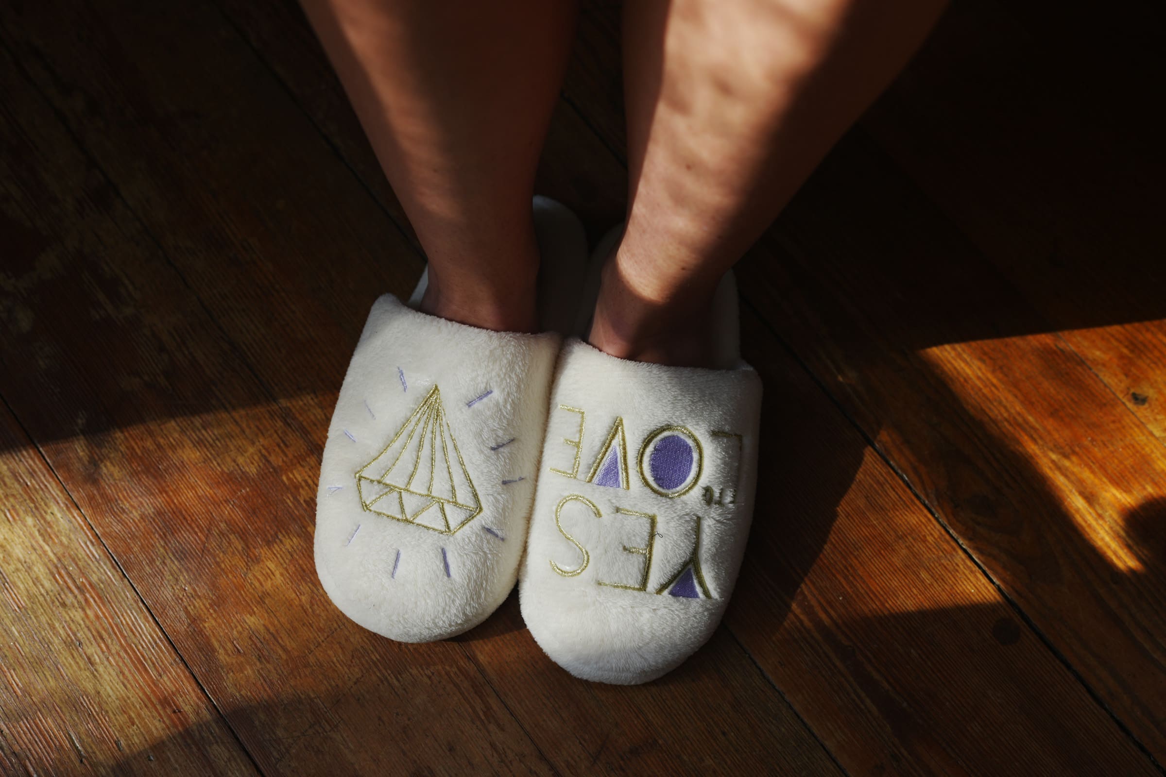 A pair of feet wearing white slippers adorned with the words YES LOVE and a diamond illustration stand gracefully on a wooden floor, illuminated by sunlight at the Rose Hill Estate wedding.