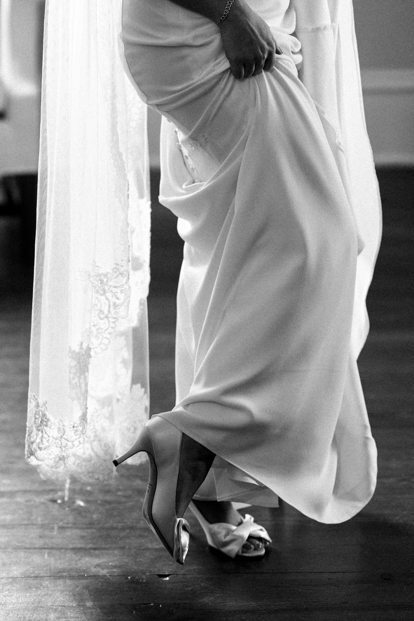 Black and white photo of a bride at Rose Hill Estate, lifting her wedding dress slightly to reveal elegant high-heeled shoes. The lace detail of the dress is visible, and the scene conveys a sense of elegance and anticipation.