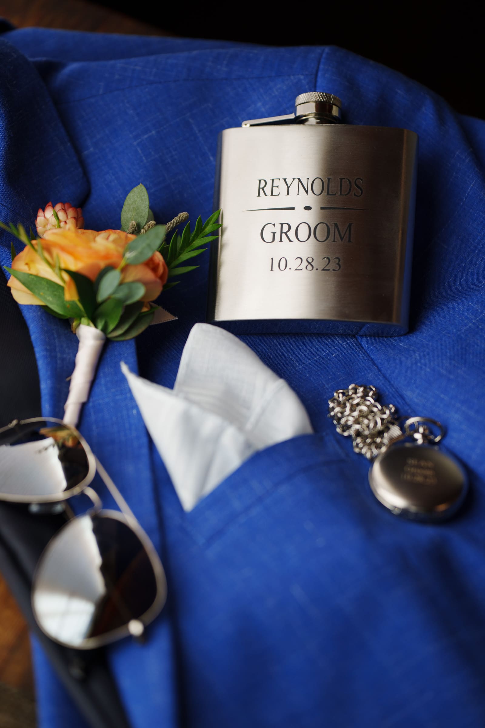 A blue suit jacket with a boutonnière featuring orange flowers, perfectly capturing the elegance of a Rose Hill Estate wedding. Beside it, a silver hip flask engraved with Reynolds Groom 10.28.23, a pocket watch, and sunglasses are all arranged neatly.